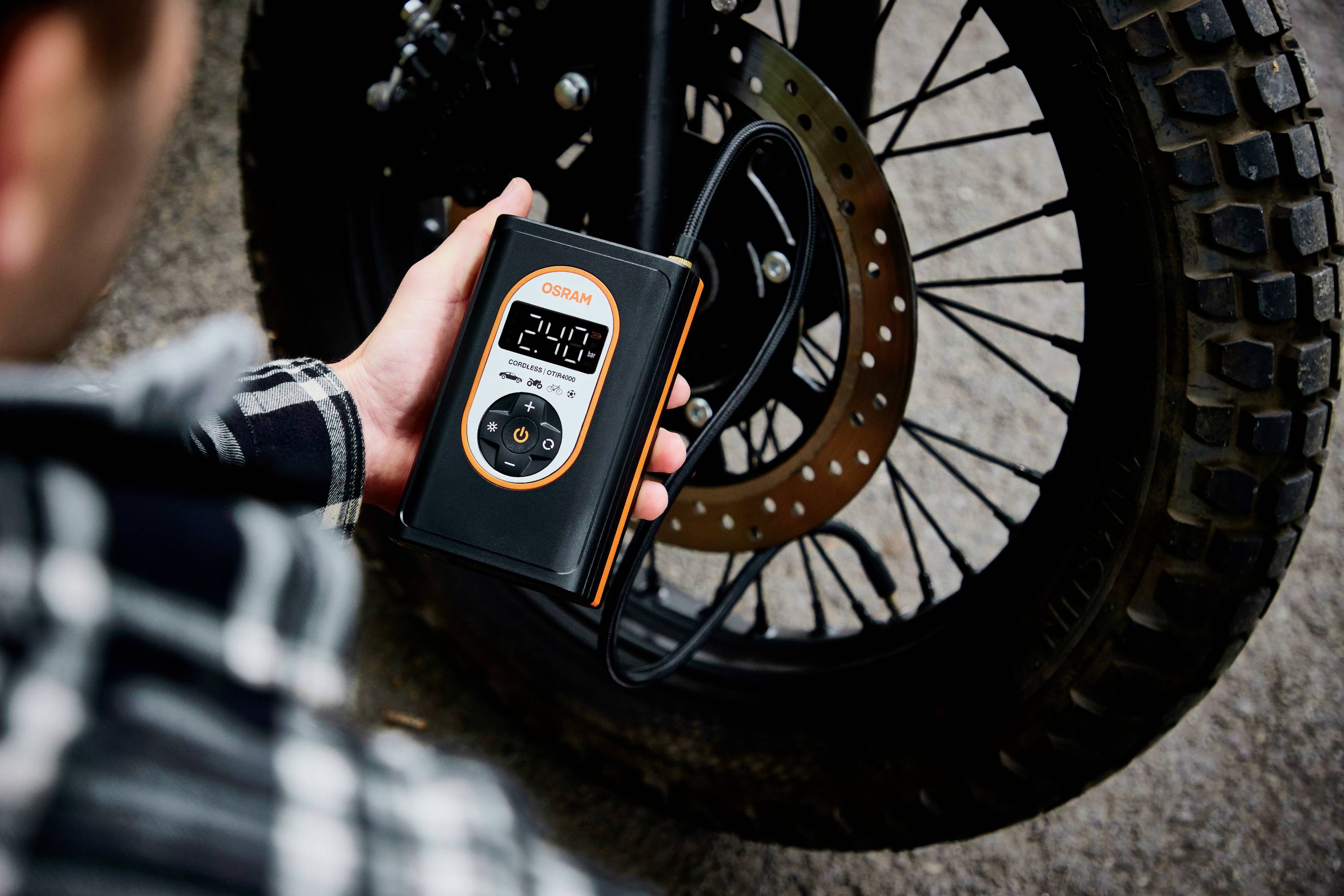 Osoba kontroluje pomocí digitálního přístroje tlak vzduchu v pneumatice motocyklu. Displej ukazuje hodnotu '2.4'.