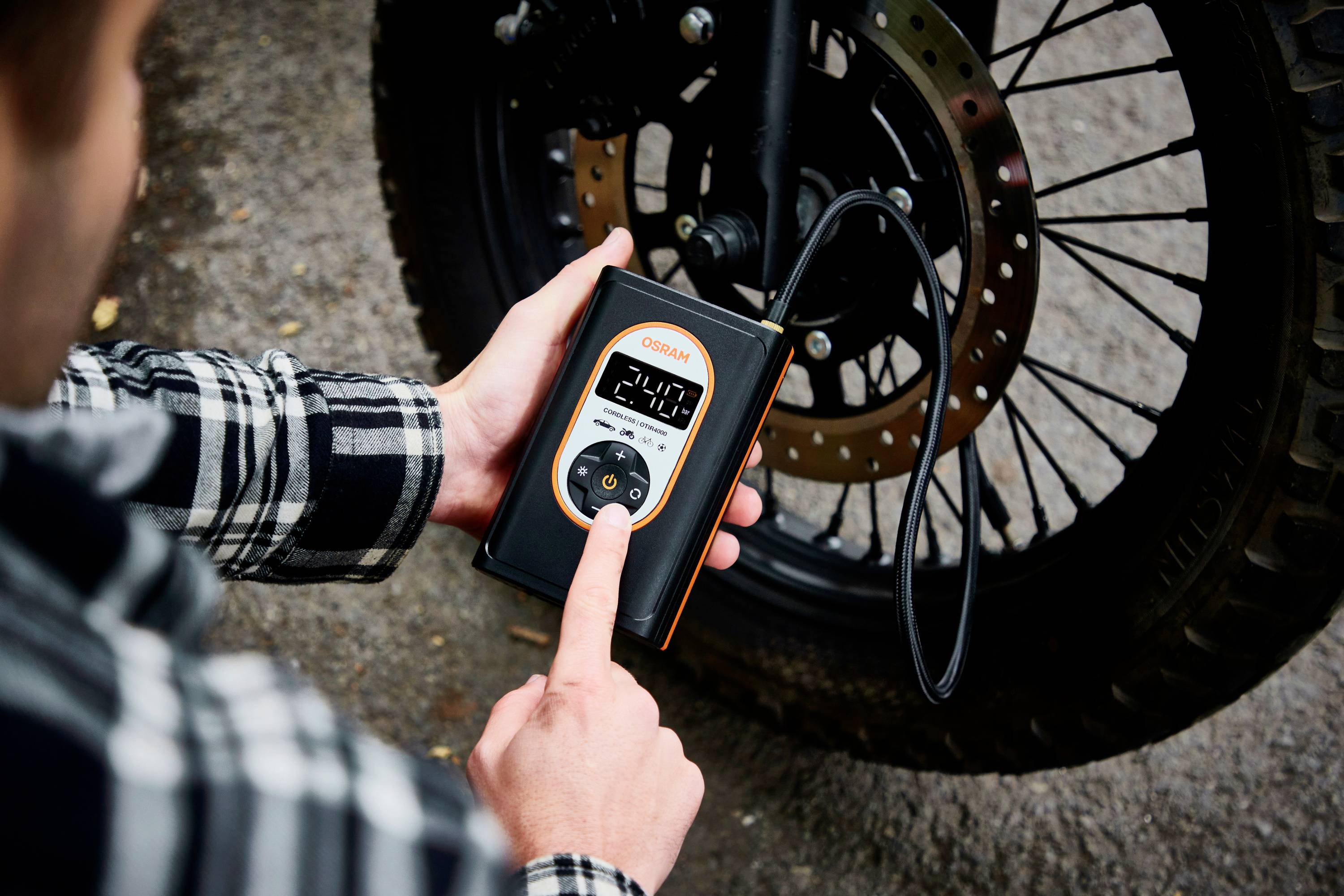 Jedna osoba kontroluje tlak vzduchu v pneumatice motocyklu pomocí digitálního tlakoměru. Displej ukazuje '2.40'.