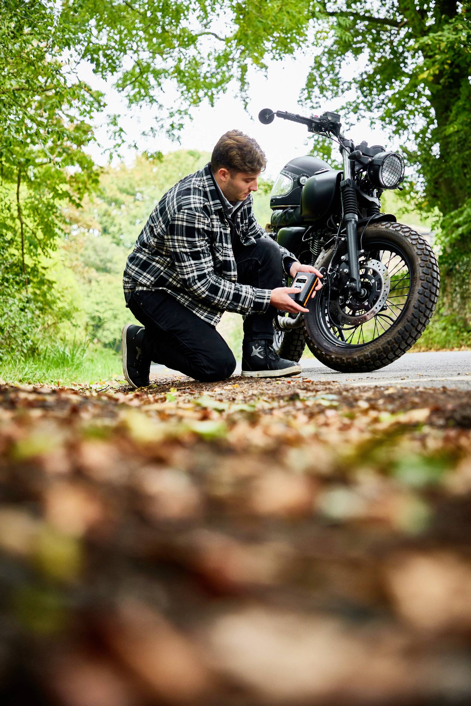 Osoba klečí na silnici a kontroluje tlak v pneumatikách motocyklu v zelené krajině. V popředí jsou viditelné podzimní listy.