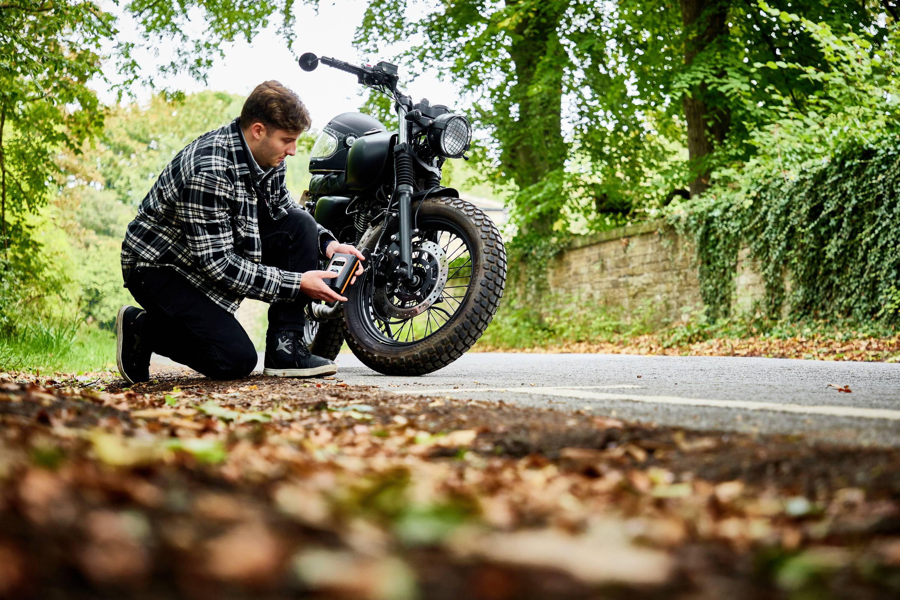 Muž klečí na silnici a pomocí měřicího přístroje kontroluje tlak v pneumatice motocyklu na lesnatém místě.