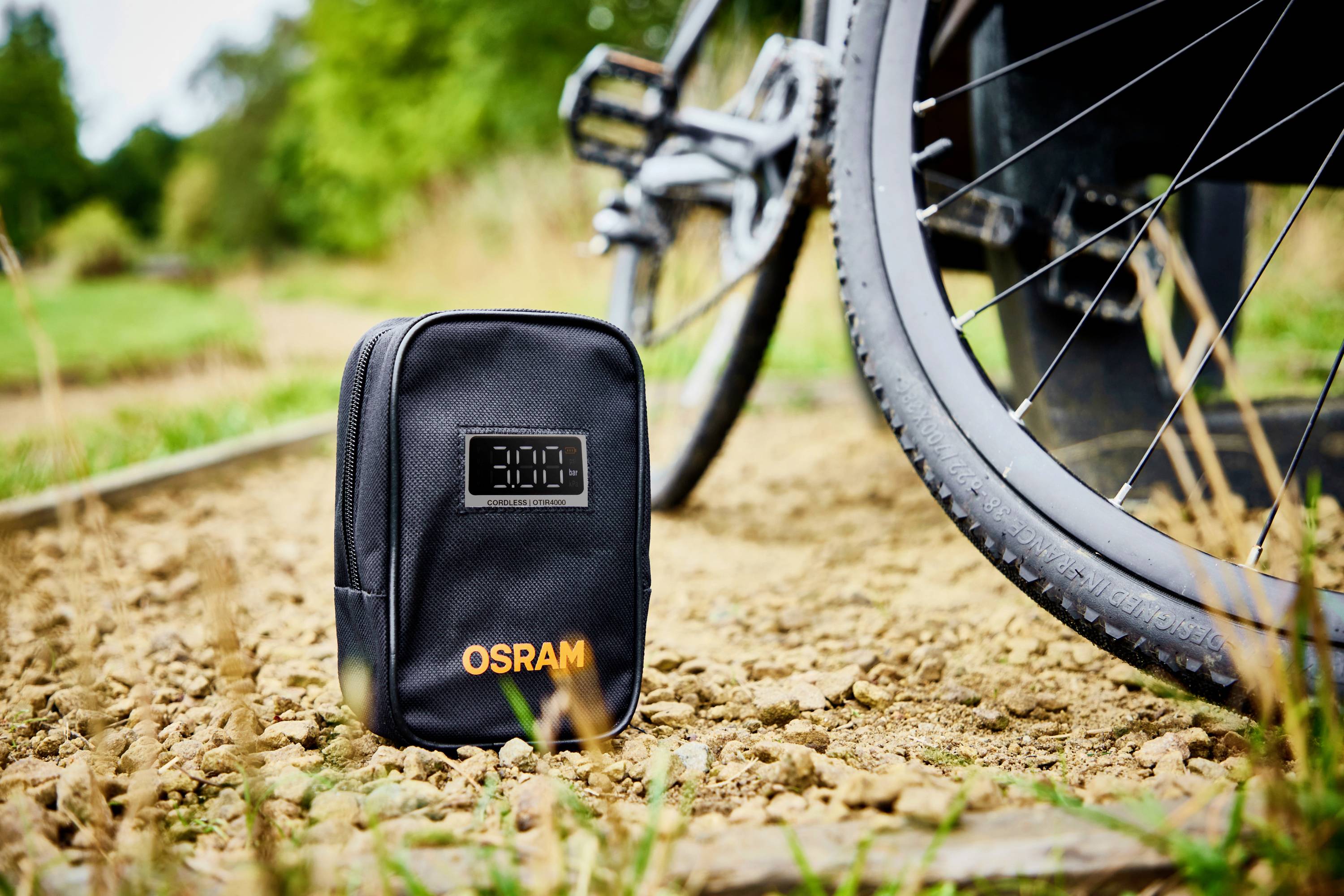 Černá taška s potiskem 'OSRAM' stojí na písčité cestě vedle cyklistické pneumatiky, v pozadí jsou viditelné stromy.