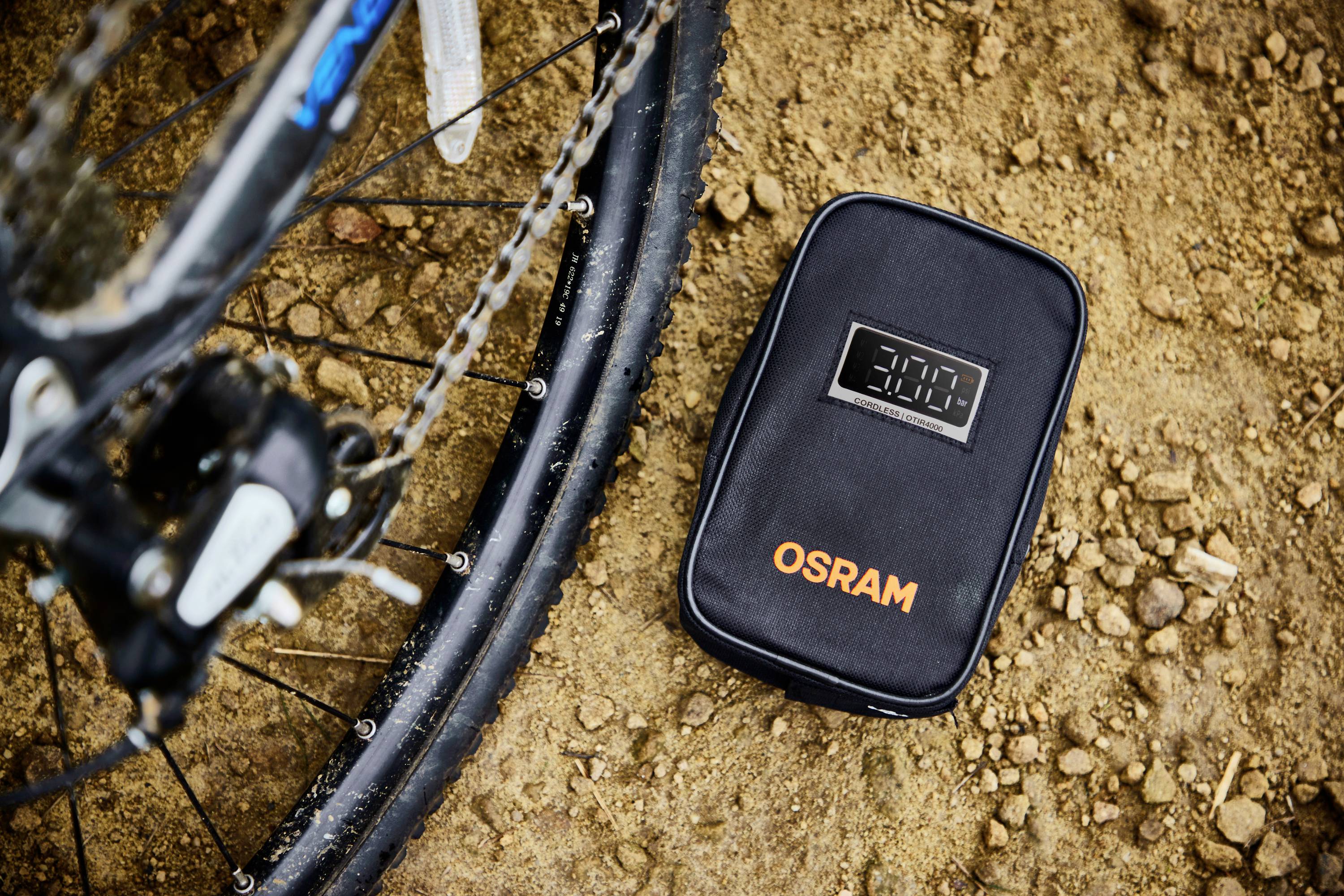 Cyklistická pneumatika a černá taška s logem 'OSRAM' a digitálním displejem leží na zemité podložce.