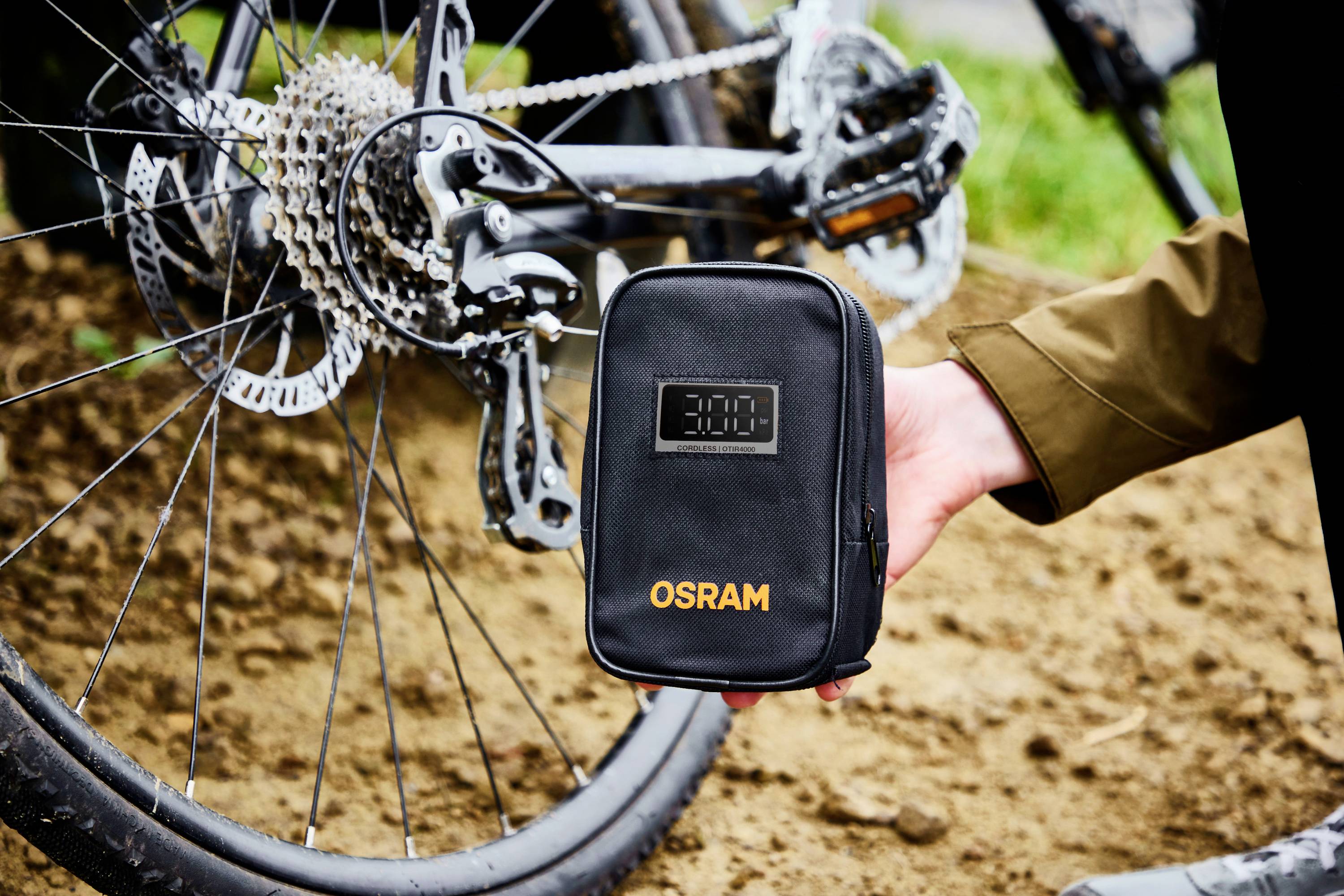 Osoba drží černou tašku s nápisem "OSRAM" vedle jízdního kola. Kolo je vybaveno ozubenými koly a kotoučovou brzdou.