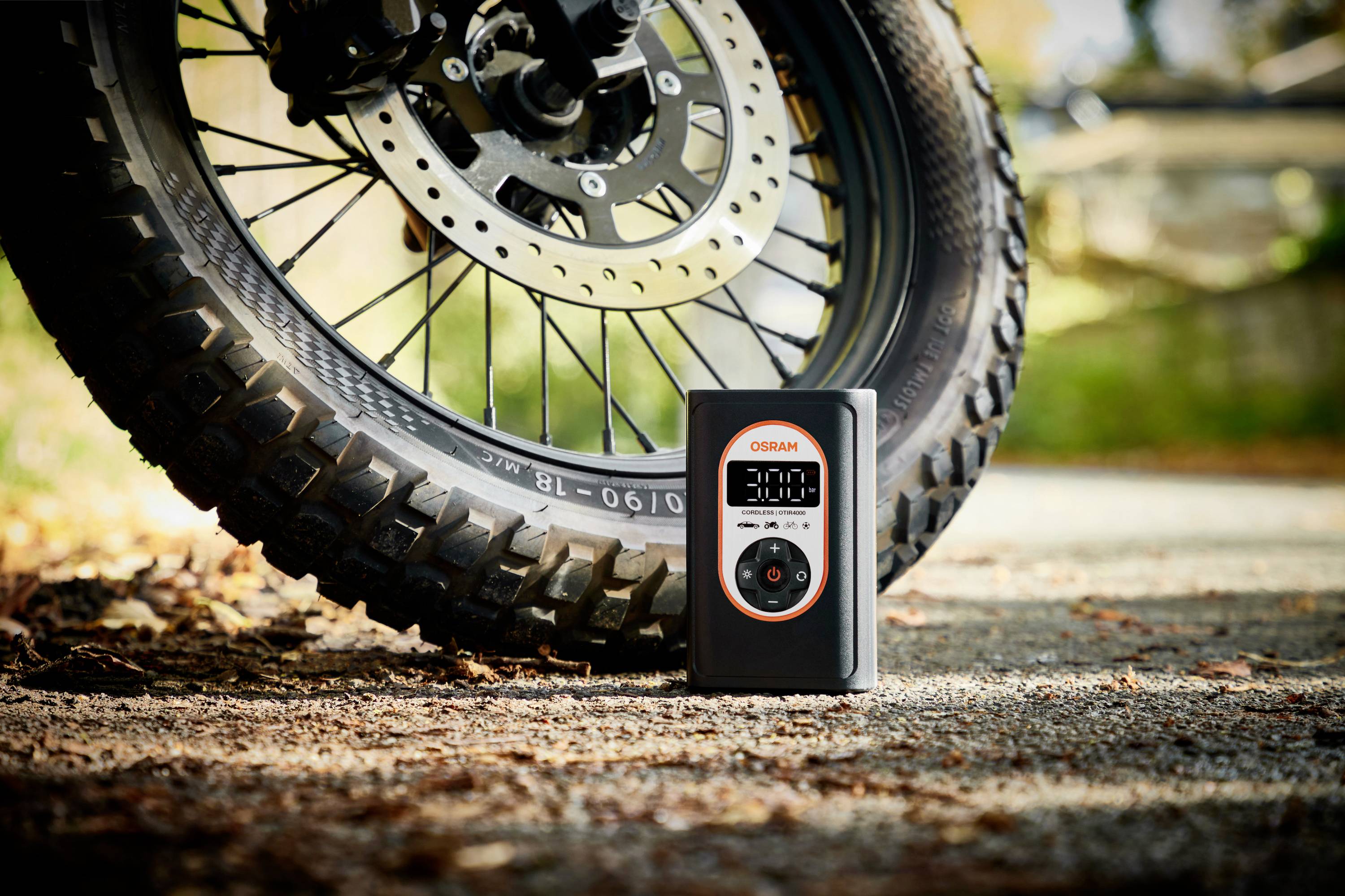Motocyklová pneumatika na asfaltu, vedle digitální měřič tlaku pneumatik od Osramu, který ukazuje 3,00 Bar. Podzimní prostředí.