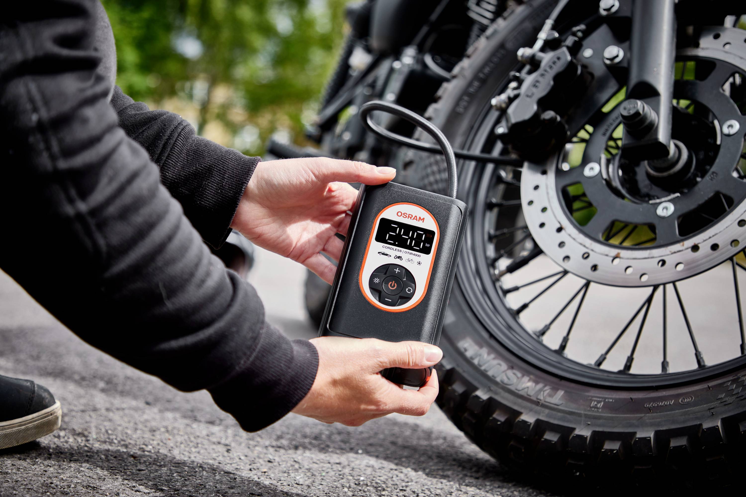 Osoba měří tlak vzduchu v pneumatice motocyklu digitálním měřicím přístrojem. V pozadí jsou viditelné stromy.
