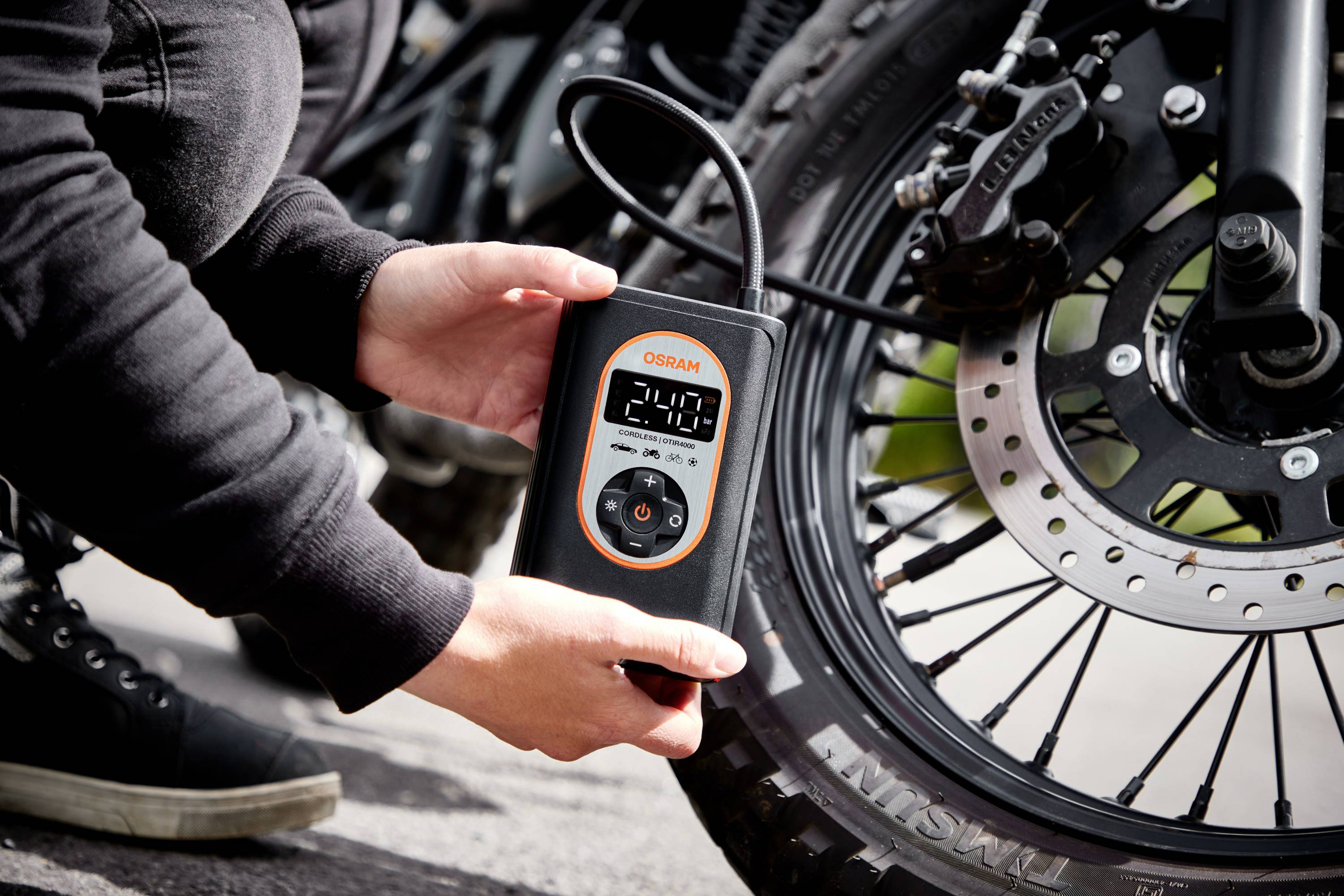 Osoba kontroluje tlak vzduchu v pneumatice motocyklu digitálním tlakoměrem, který ukazuje 2,4 baru.