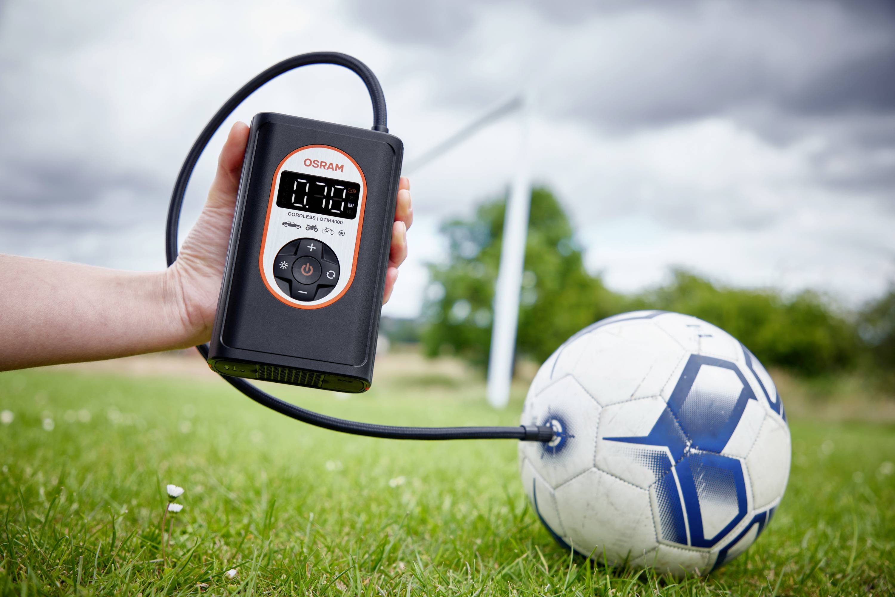 Jedna ruka drží elektrickou vzduchovou pumpu, která nafukuje fotbalový míč na hřišti. V pozadí je vidět brána.