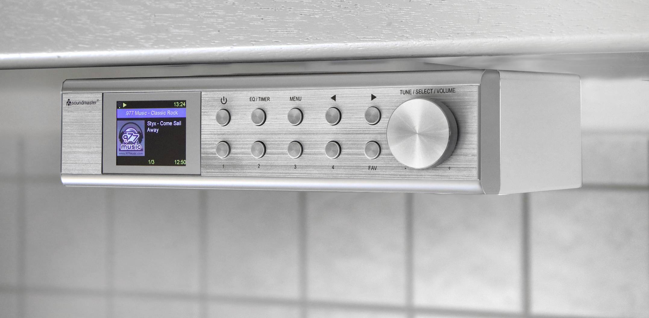 Stříbrné kuchyňské rádio s displejem a ovládacími tlačítky. Na obrazovce zobrazuje nápis 'Stop, Come Sail Away'. Upevněné pod kuchyňskou skříňkou.