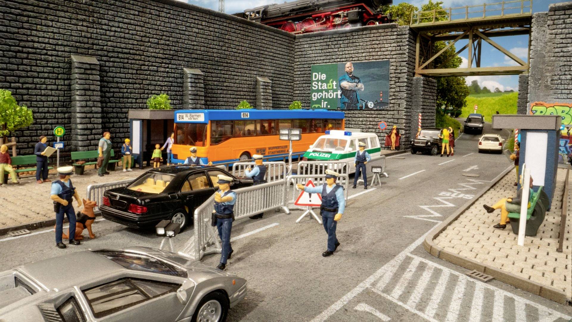 Pouliční scéna s policejní kontrolou: Autobus a auta před zátarasy, policisté a osoby vedoucí rozhovor, velký plakát v pozadí.