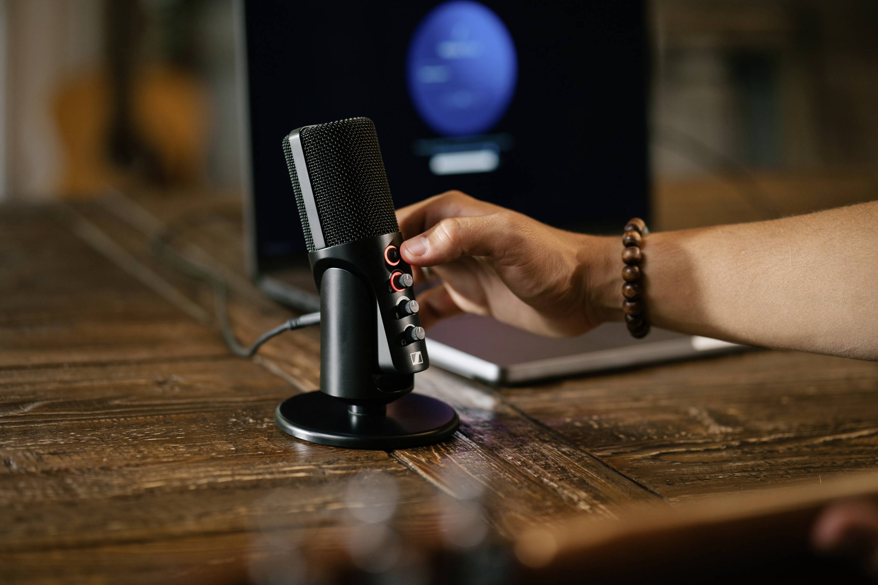 Ruka upravuje nastavení černého mikrofonu, který stojí před otevřeným laptopem na dřevěném stole.