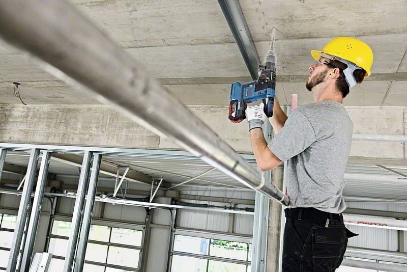 Dělník v ochranné přilbě s vrtačkou upevňuje kovový rám ke stropu v nedokončené budově.