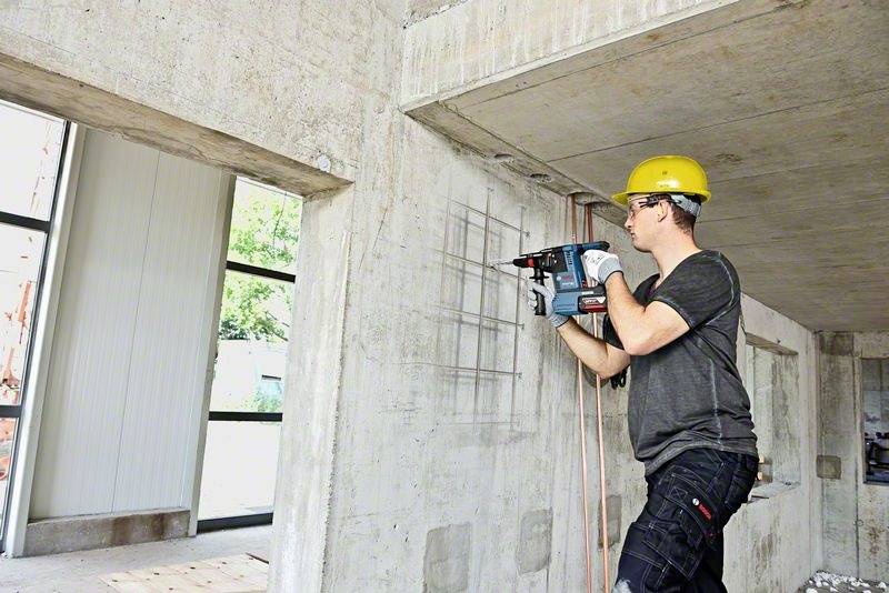 Dělník v žluté helmě vrtá pomocí vrtacího kladiva do betonové stěny v nedokončené budově.