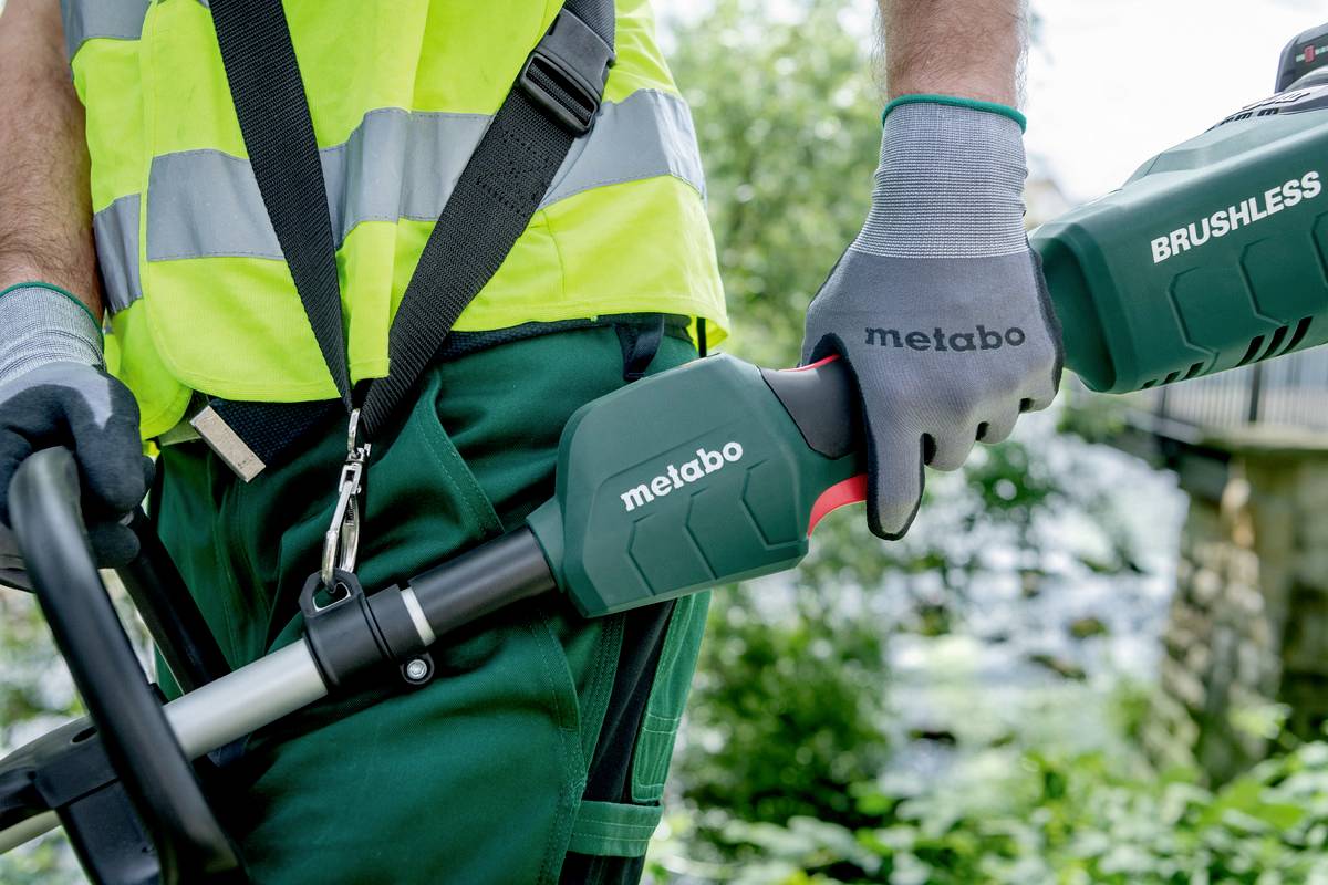 Dělník v pracovním oblečení a rukavicích drží křovinořez značky Metabo. V pozadí je viditelná zelená vegetace.