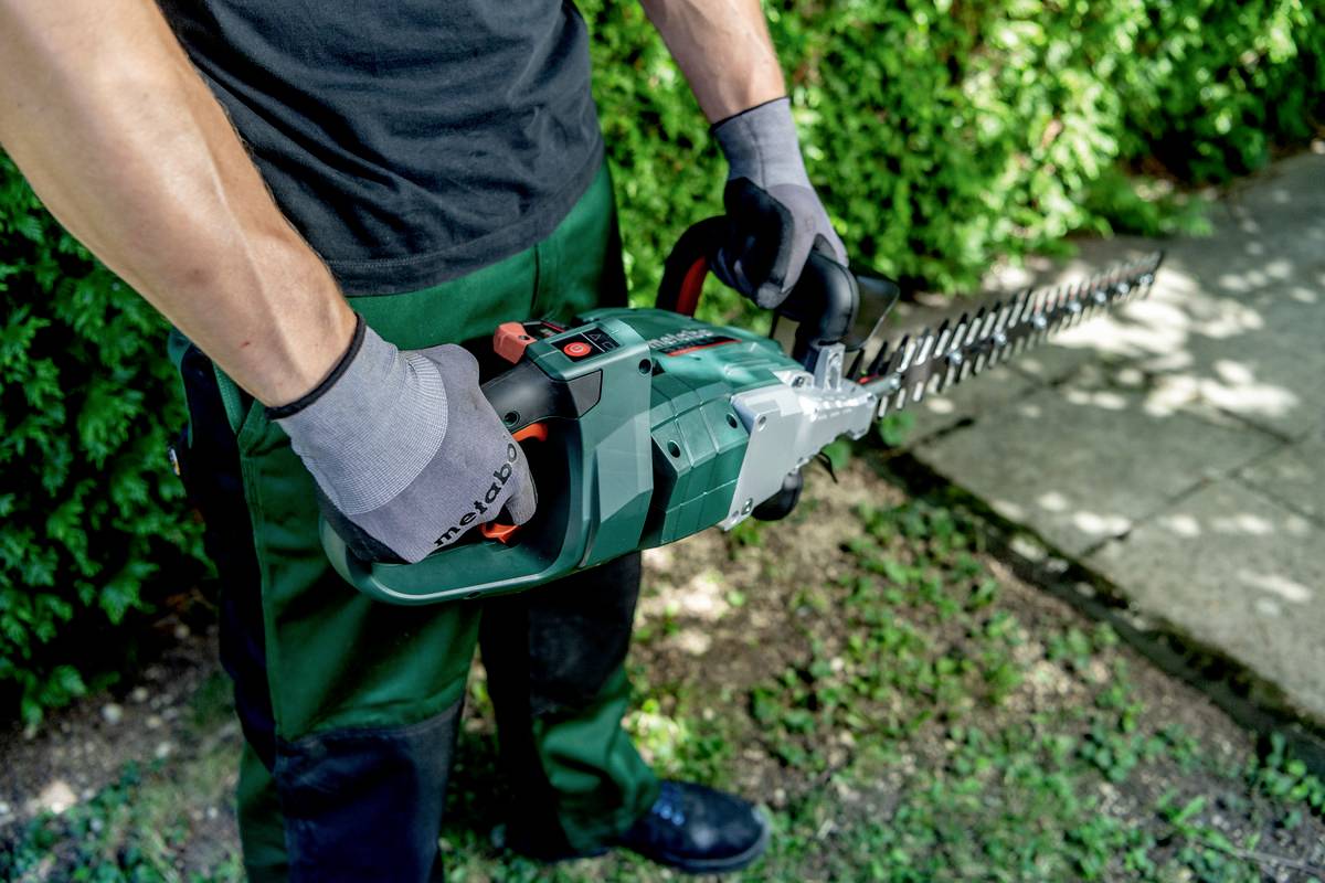 Osoba v rukavicích drží elektrické zahradní nůžky a střihá živý plot na zahradě.