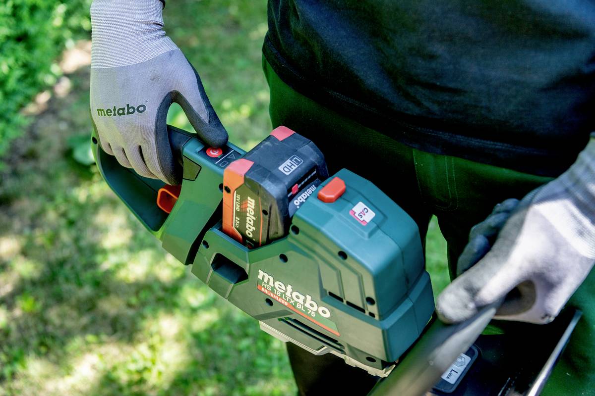 Osoba v pracovních rukavicích drží zelenou elektrickou pilu značky Metabo, která je používána v zahradě.