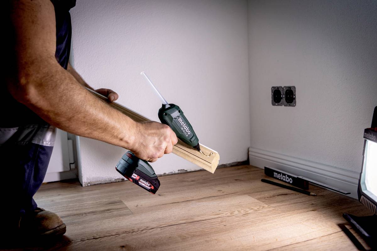 Osoba drží elektrický nástroj a kus dřeva na dřevěné podlaze; vedle je zdroj světla a elektrické zásuvky.