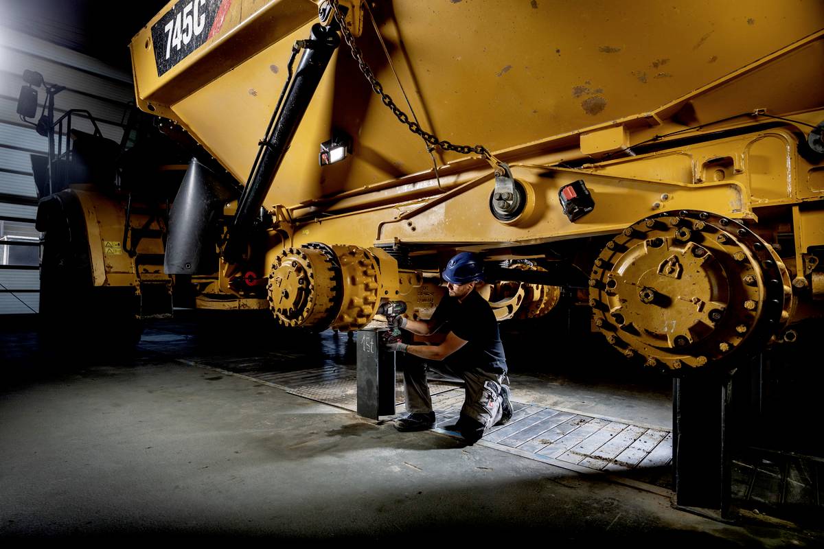 Technik pracuje na velkém žlutém průmyslovém strojním vozidle v dílně. Má na sobě ochrannou přilbu a používá nářadí.