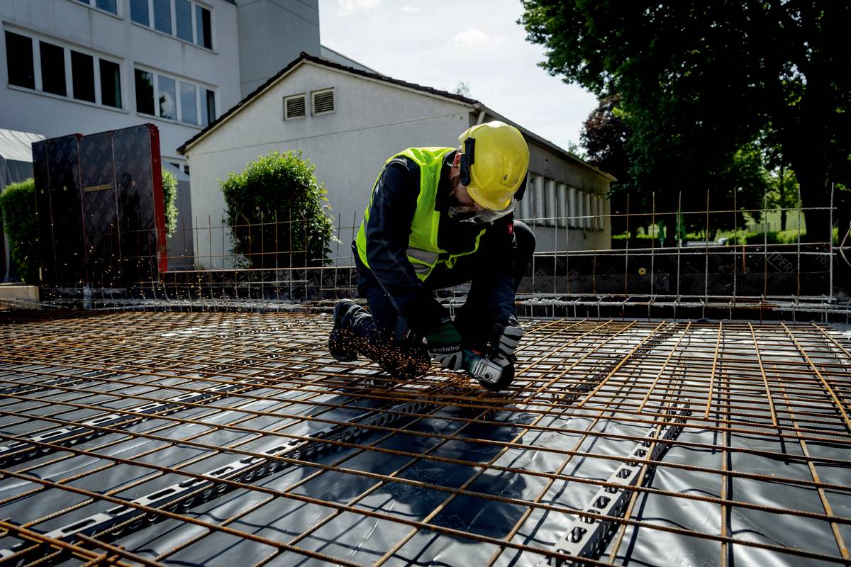 Dělník řeže kovové tyče na staveništi. Má na sobě žlutou ochrannou přilbu a zelenou bezpečnostní vestu.