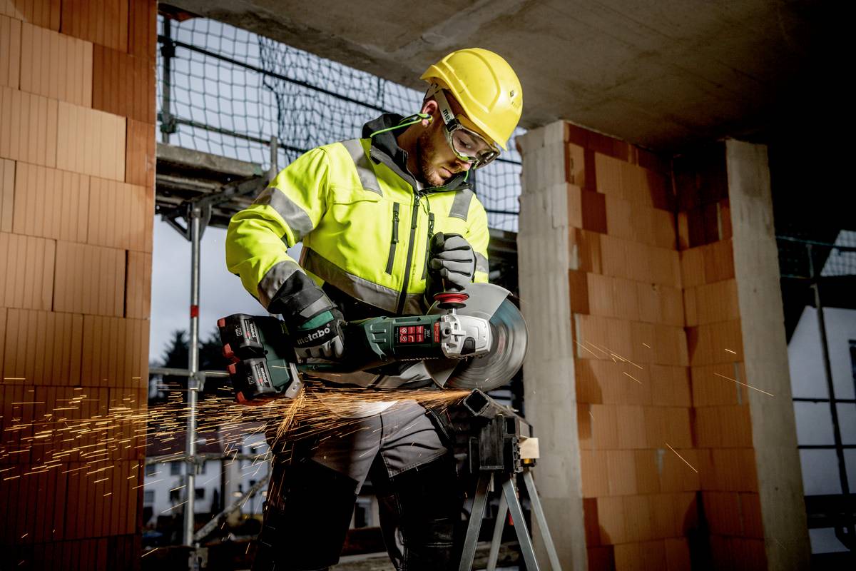 Dělník v ochranném oděvu řeže úhlovou bruskou beton, přičemž kolem létají jiskry. V pozadí je staveniště.