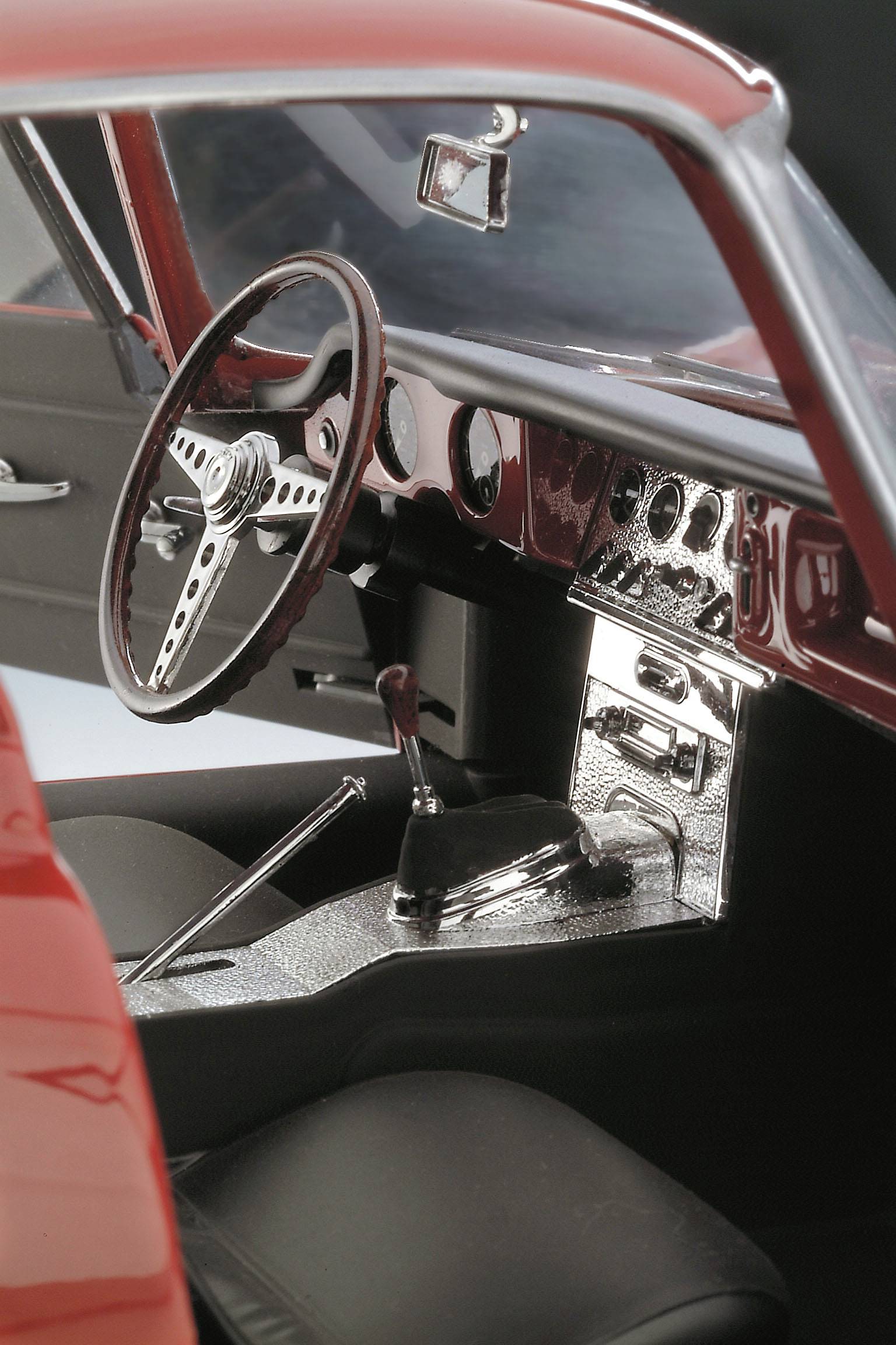 Vnitřní prostor červeného automobilu, pohled na volant a přístrojovou desku s různými tlačítky a pákami, černé sedačky, sportovní design.
