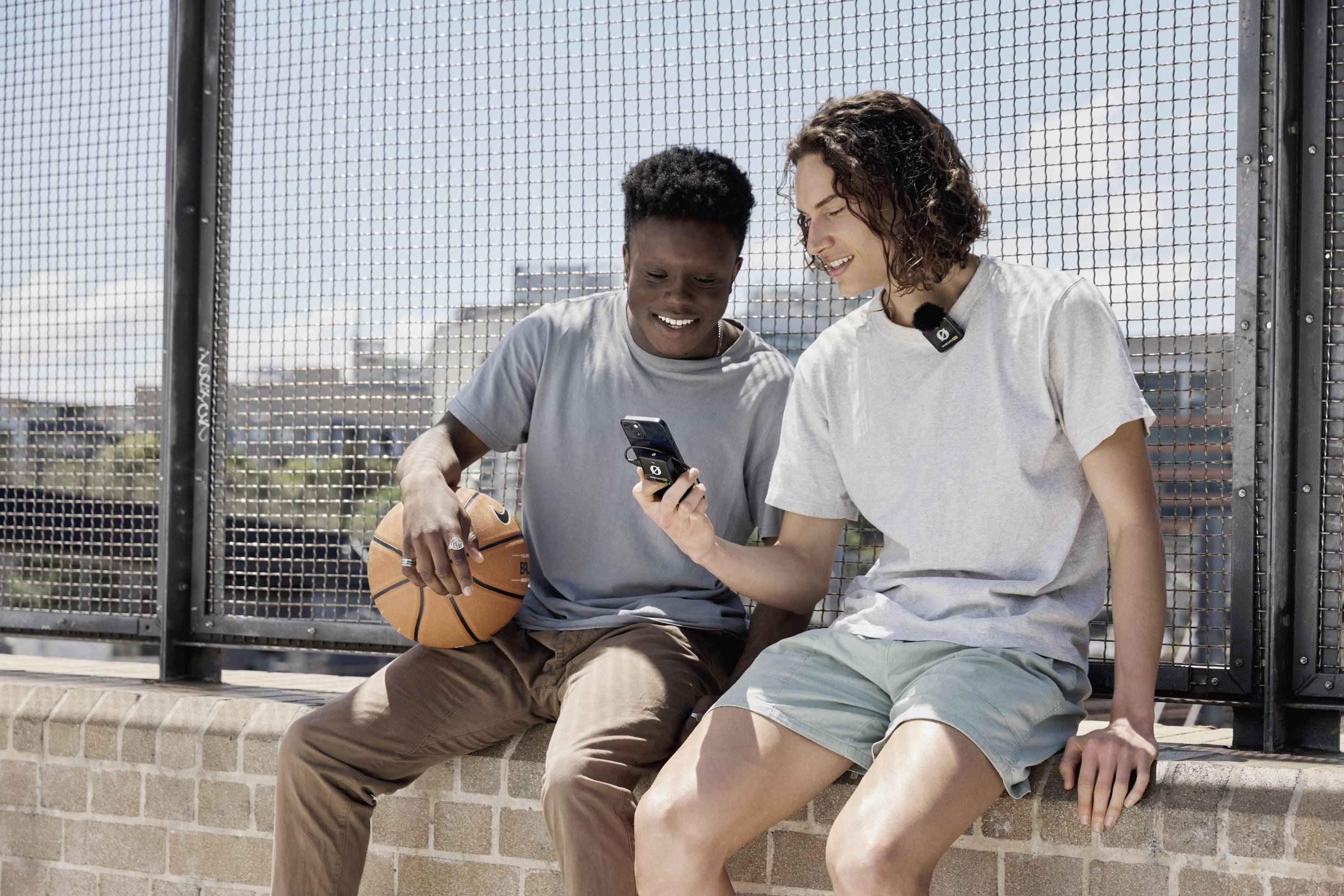 Dva mladí muži sedí na zdi. Jeden drží basketbalový míč, druhý mu ukazuje něco na smartphonu. Působí uvolněně.