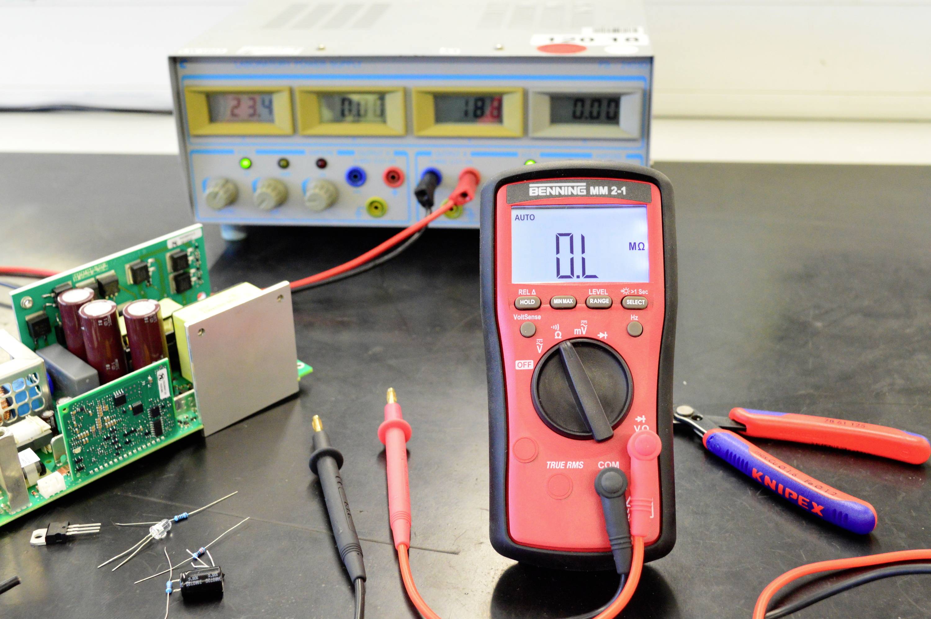 Digitální multimetr ukazuje nulu ohmů, vedle něj jsou elektronické komponenty a napájecí zdroj s osvětleným displejem.