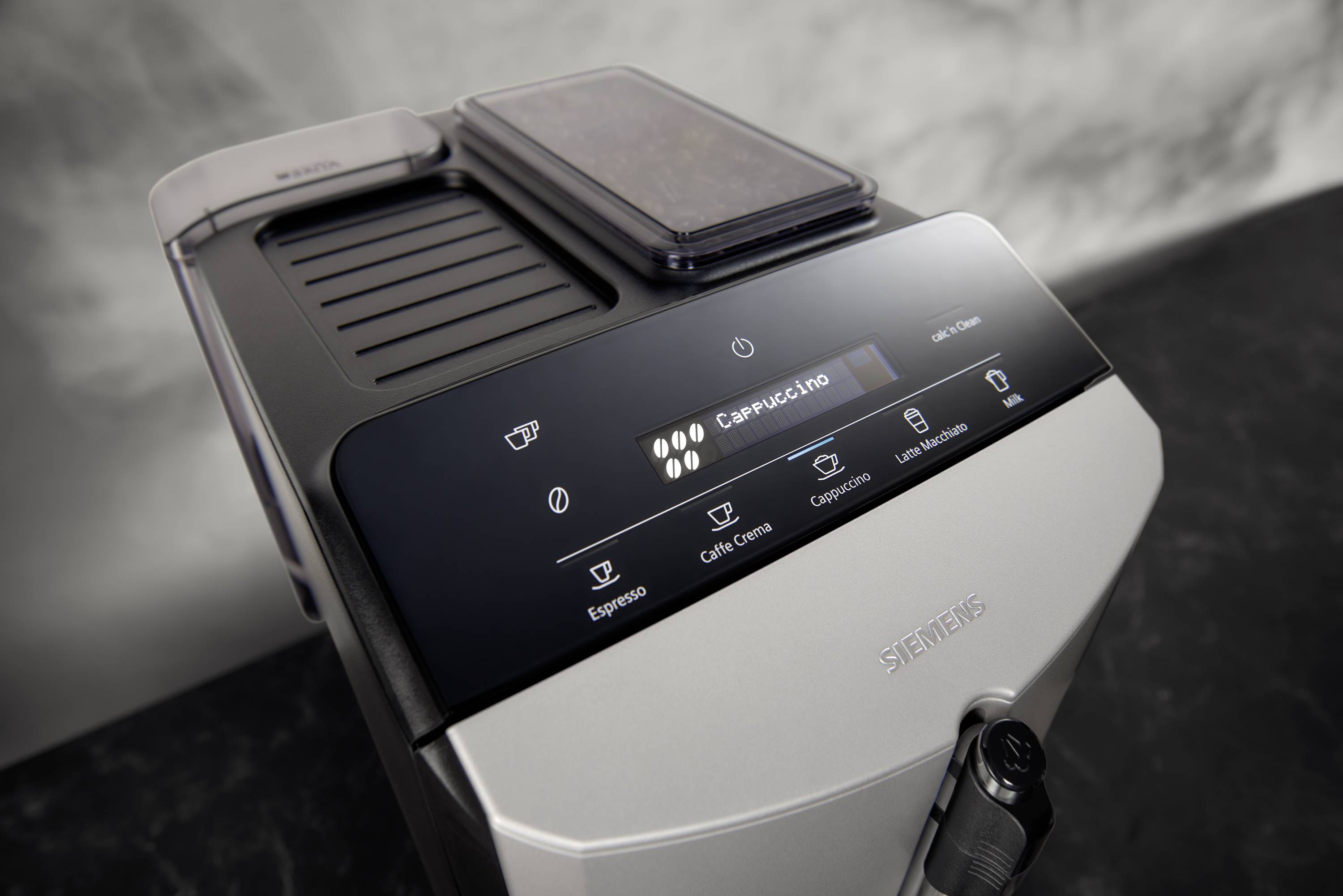 Kávovací přístroj s digitálním displejem, zobrazující 'Cappuccino'. Symboly pro velikost šálku a sílu kávy. Černé a stříbrné provedení přístroje.