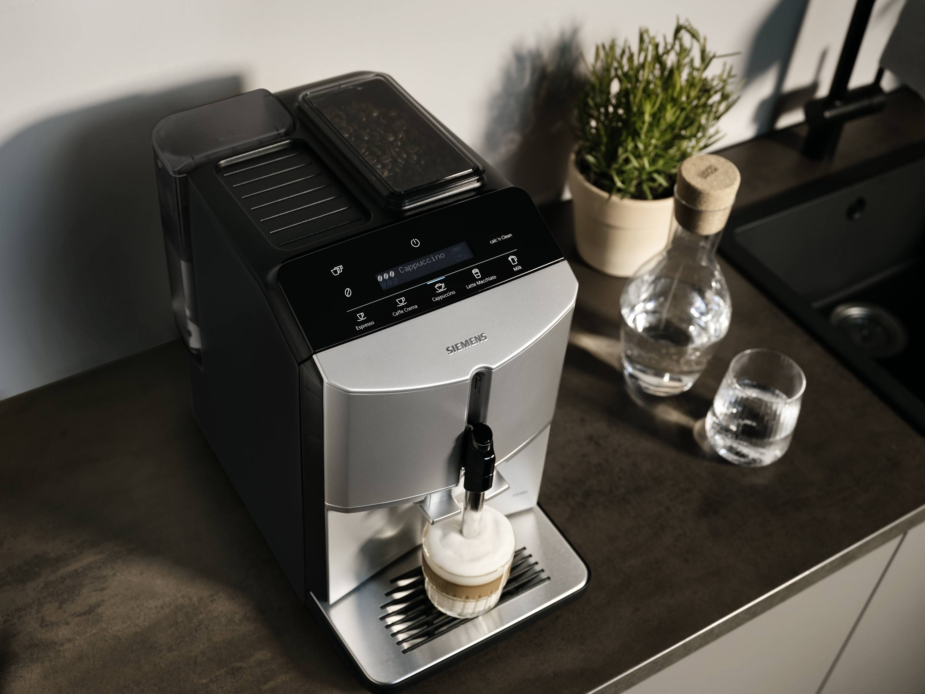 Plně automatický kávovar na kuchyňské lince připravuje latté macchiato; vedle něj stojí džbán vody a sklenice.