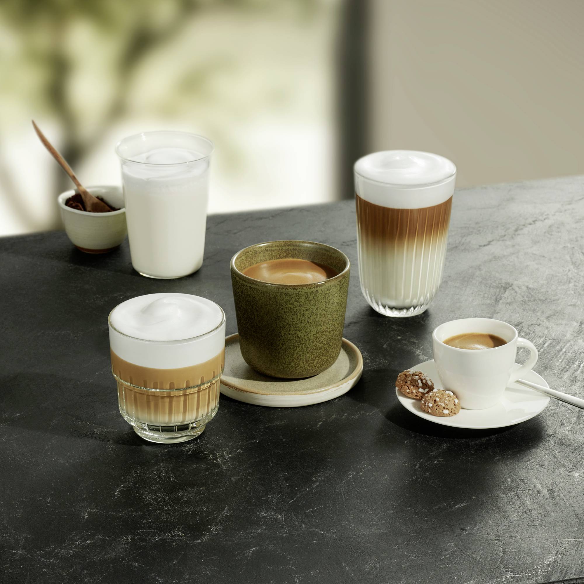 Čtyři šálky kávy s různými druhy nápojů na stole: cappuccino, latte macchiato, café au lait a espresso s pečivem.