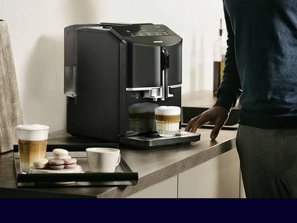 Osoba obsluhuje kávovar, který právě připravuje cappuccino. Na stole stojí šálky s kávou a sušenky.