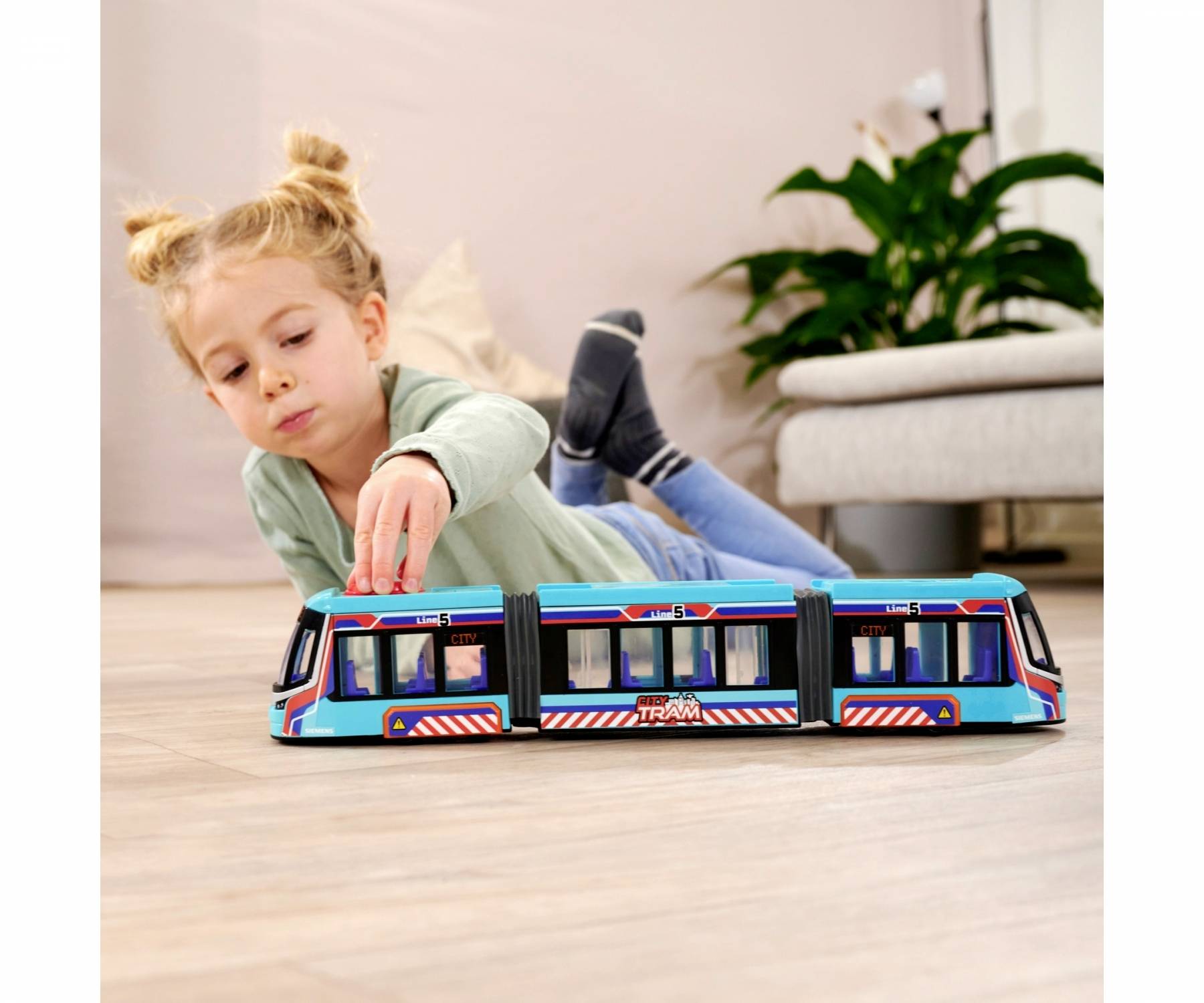 Dítě si hraje na podlaze s modrou modelovou tramvají v útulném obývacím pokoji s rostlinou v pozadí.
