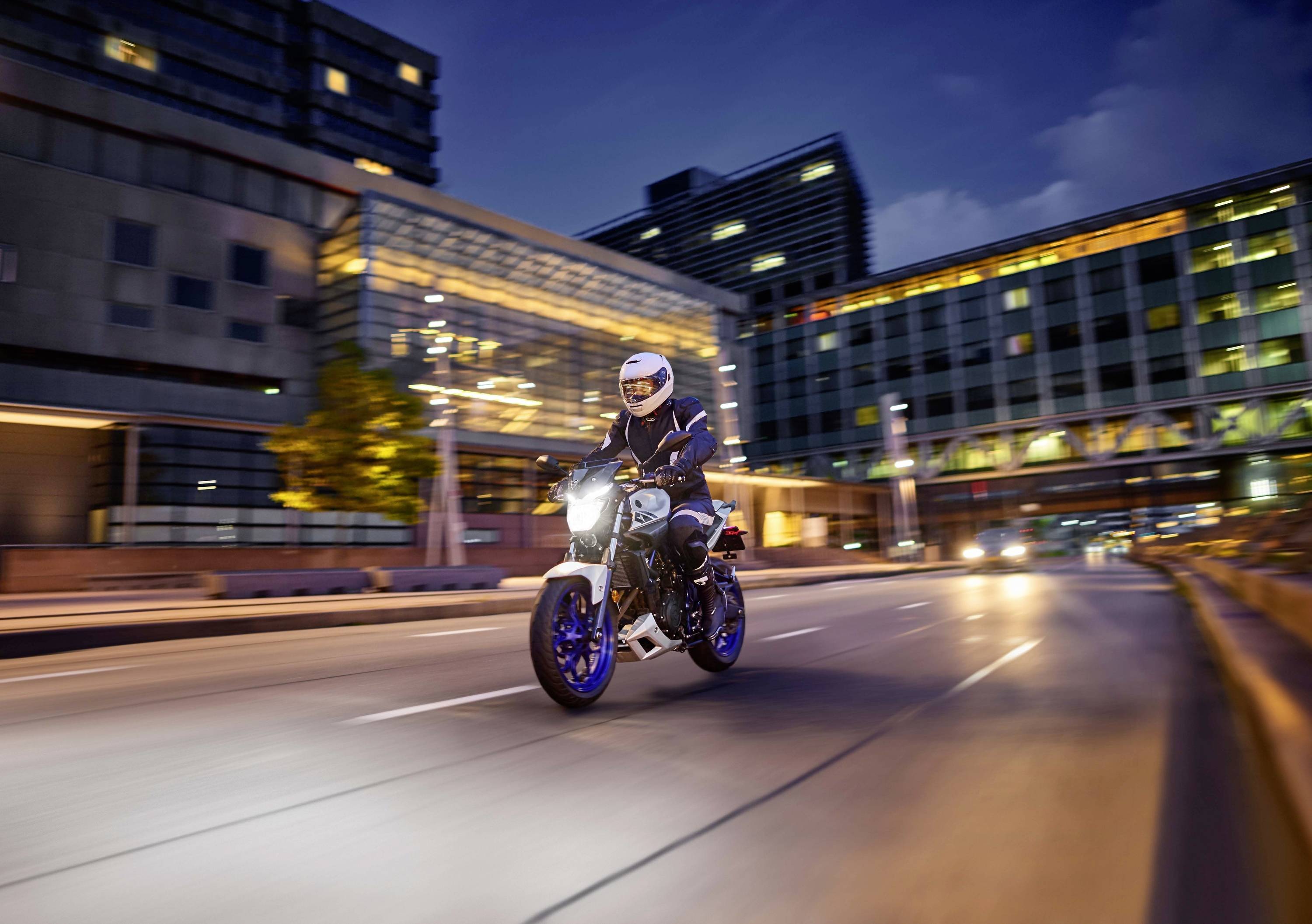 Osoba jede na osvětleném motocyklu za soumraku prázdnou městskou ulicí, obklopena moderními budovami.