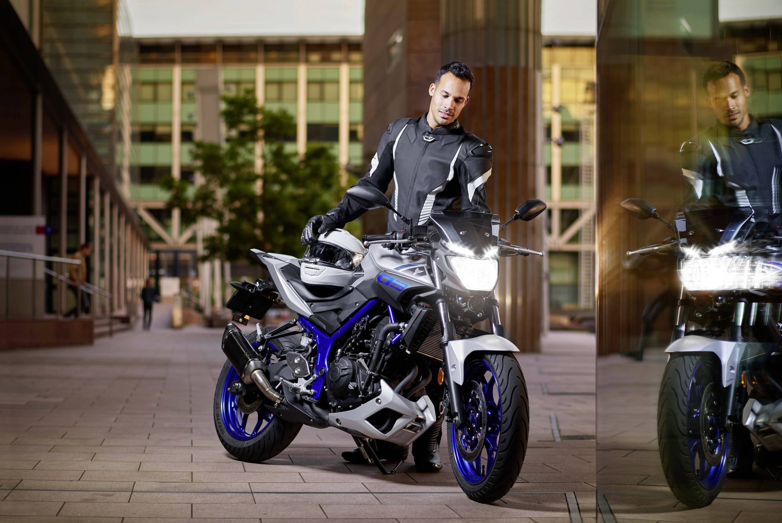 Člověk stojí vedle sportovní motorky, je oblečen v ochranném oblečení. Odrazy ve skle. Městská krajina v pozadí.