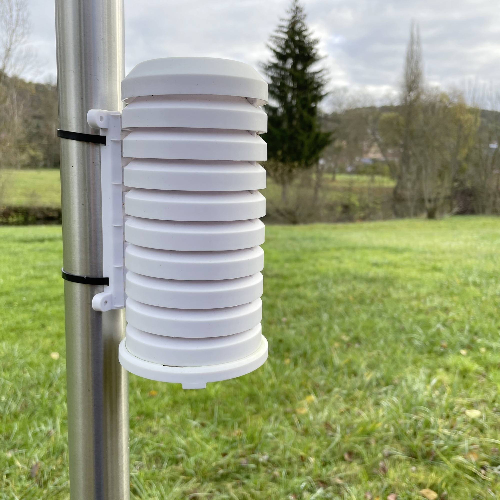 Bílý meteorologický senzor upevněný na kovovém sloupu měří klimatické údaje. V pozadí jsou viditelné travnatá plocha a stromy.