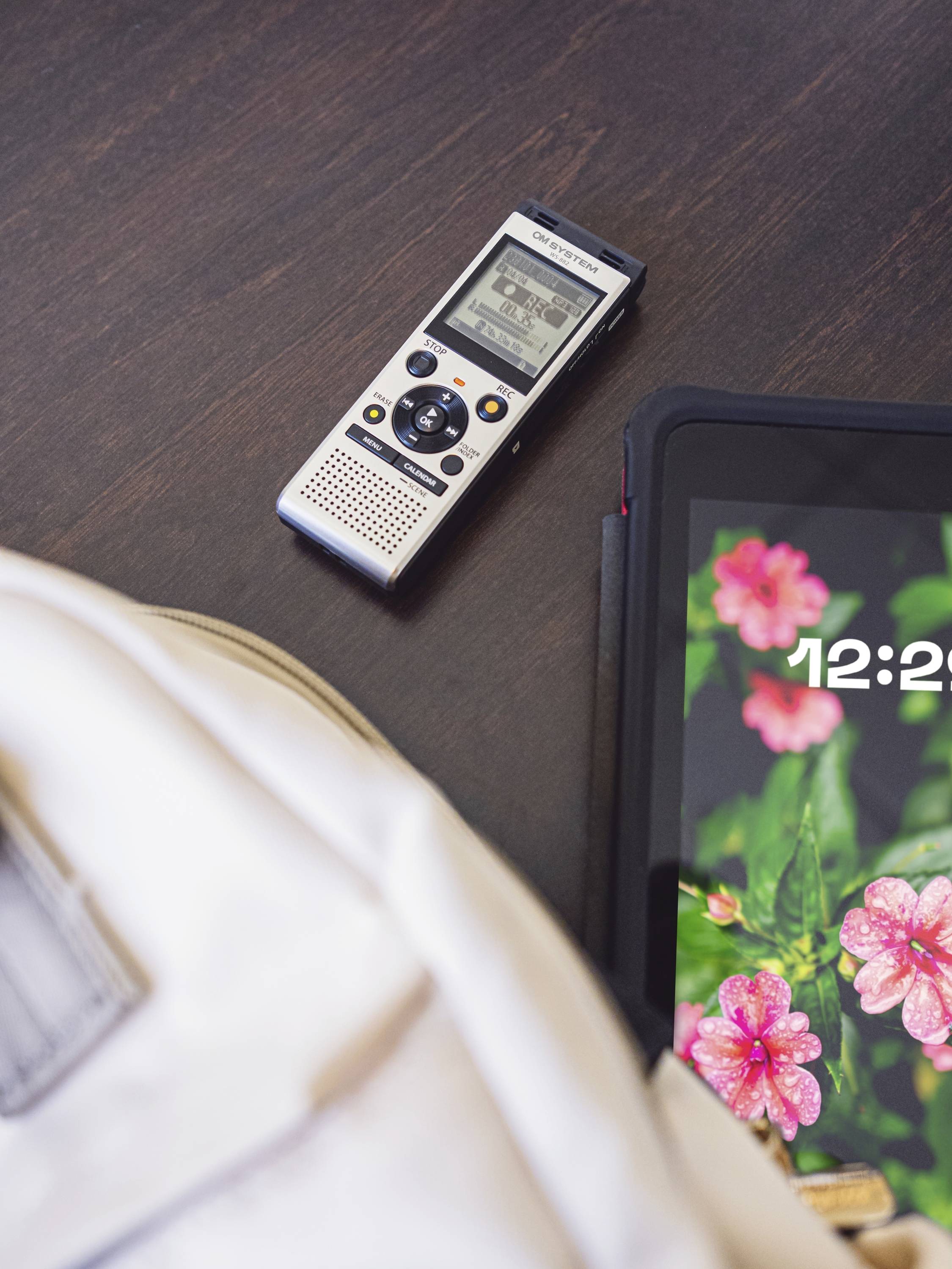 Černý stůl s digitálním diktafonem a tabletem. Tablet zobrazuje čas '12:22' a motiv květiny.