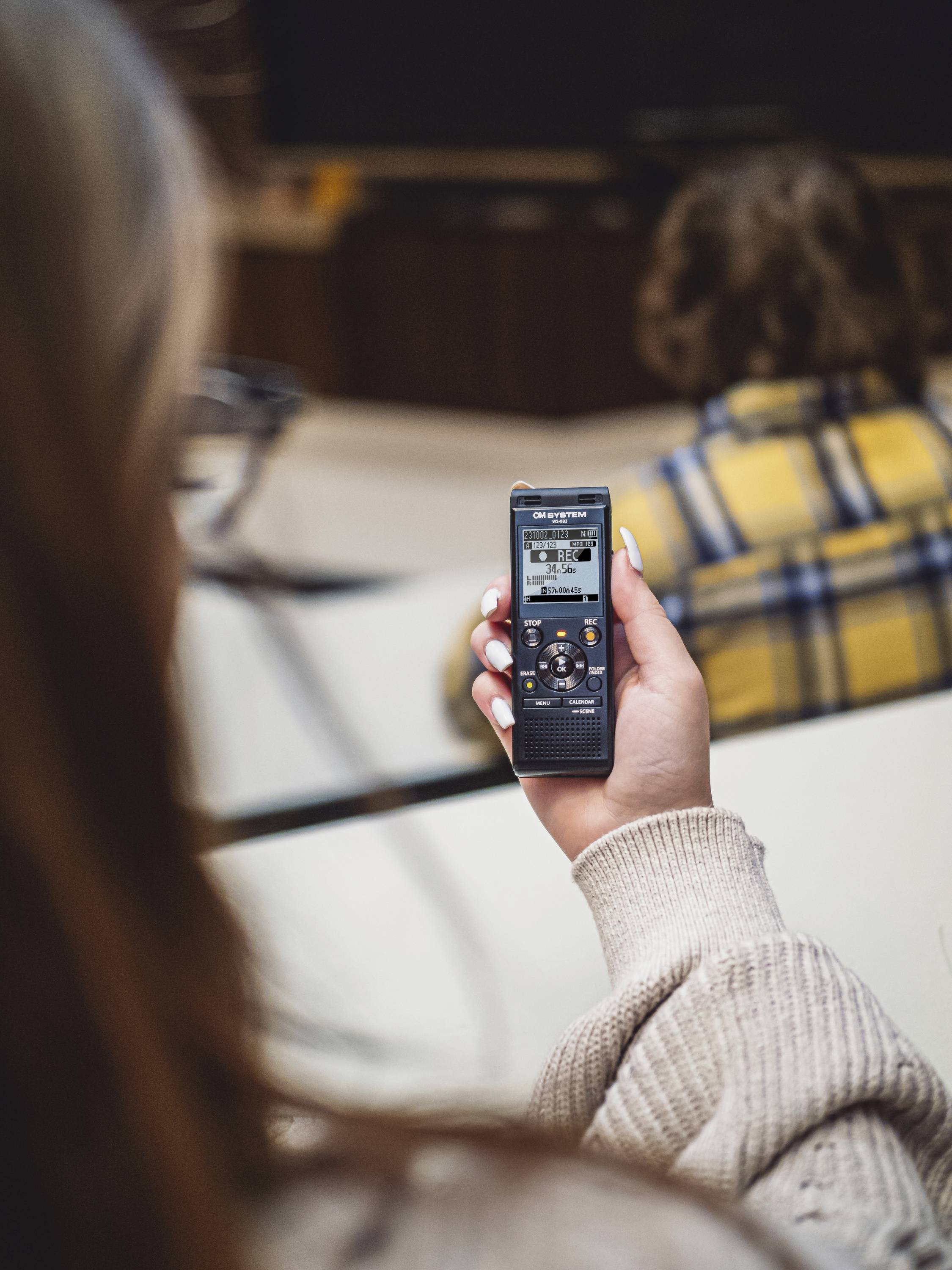Jedna osoba drží digitální audiorekordér v ruce. V pozadí sedí další rozmazaná osoba u stolu.