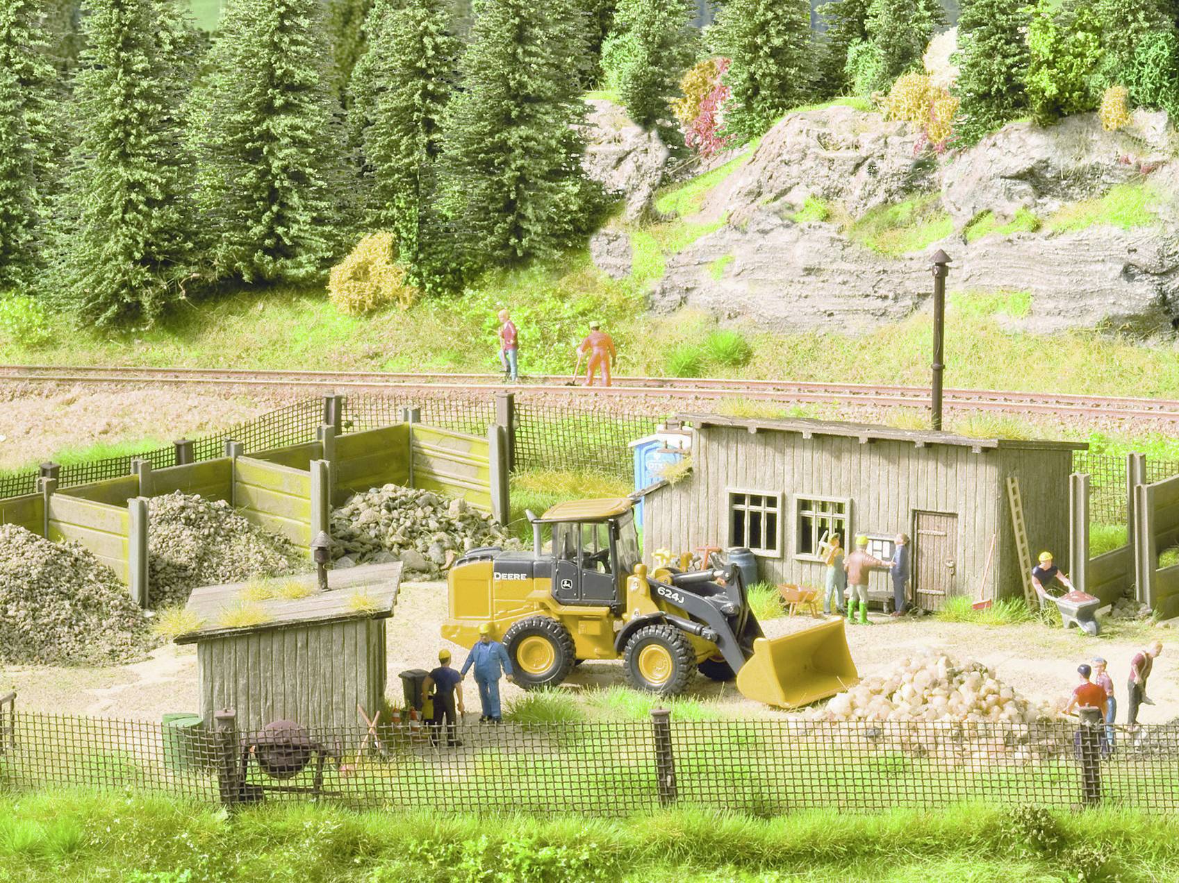 Model diorama zobrazuje staveniště s bagrem, dělníky, dřevěnou chatkou a okolními stromy a skálami.