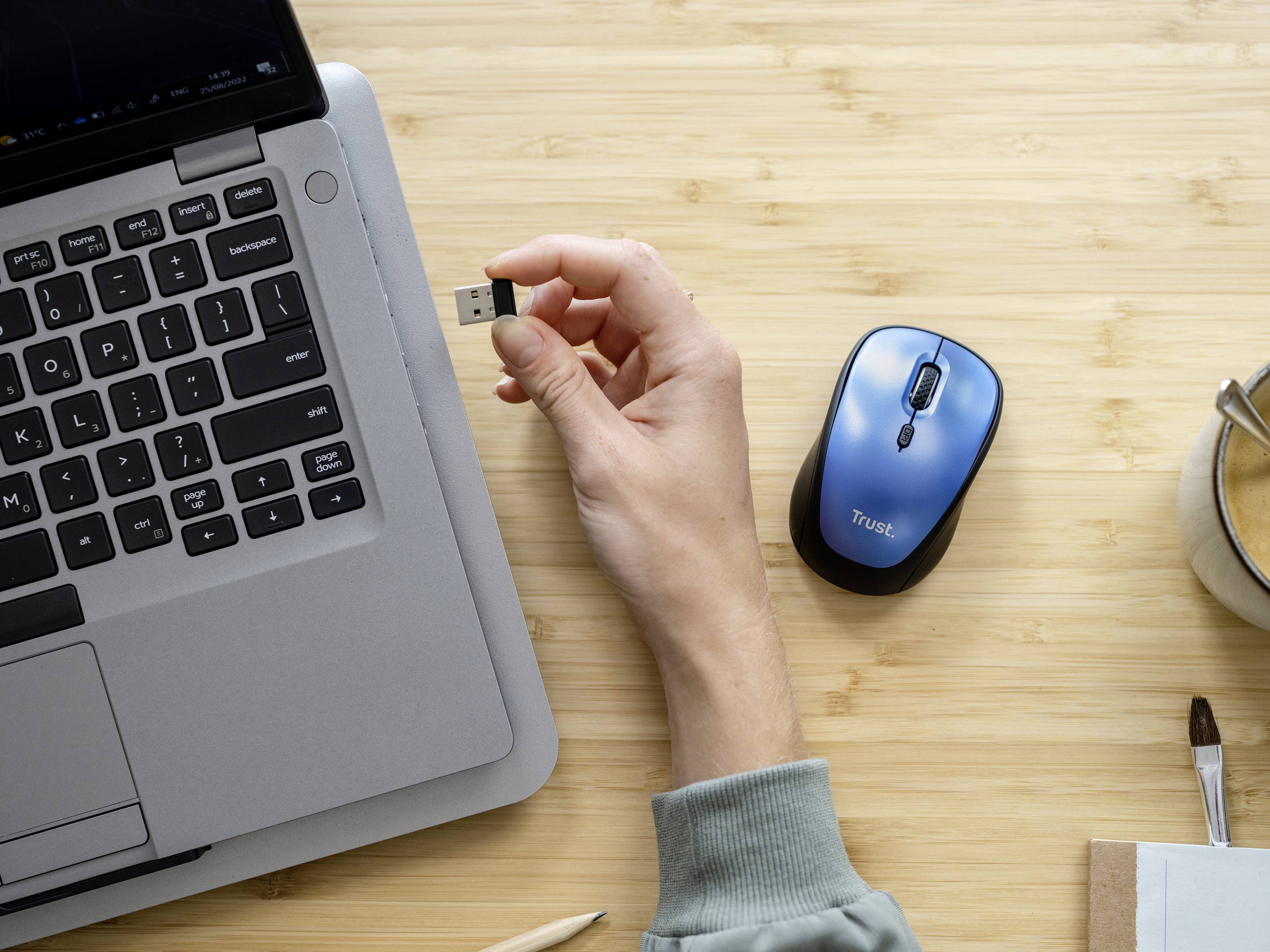 Ruka zasunuje USB flash disk do laptopového počítače, vedle které leží modrá bezdrátová myš na dřevěném stole.