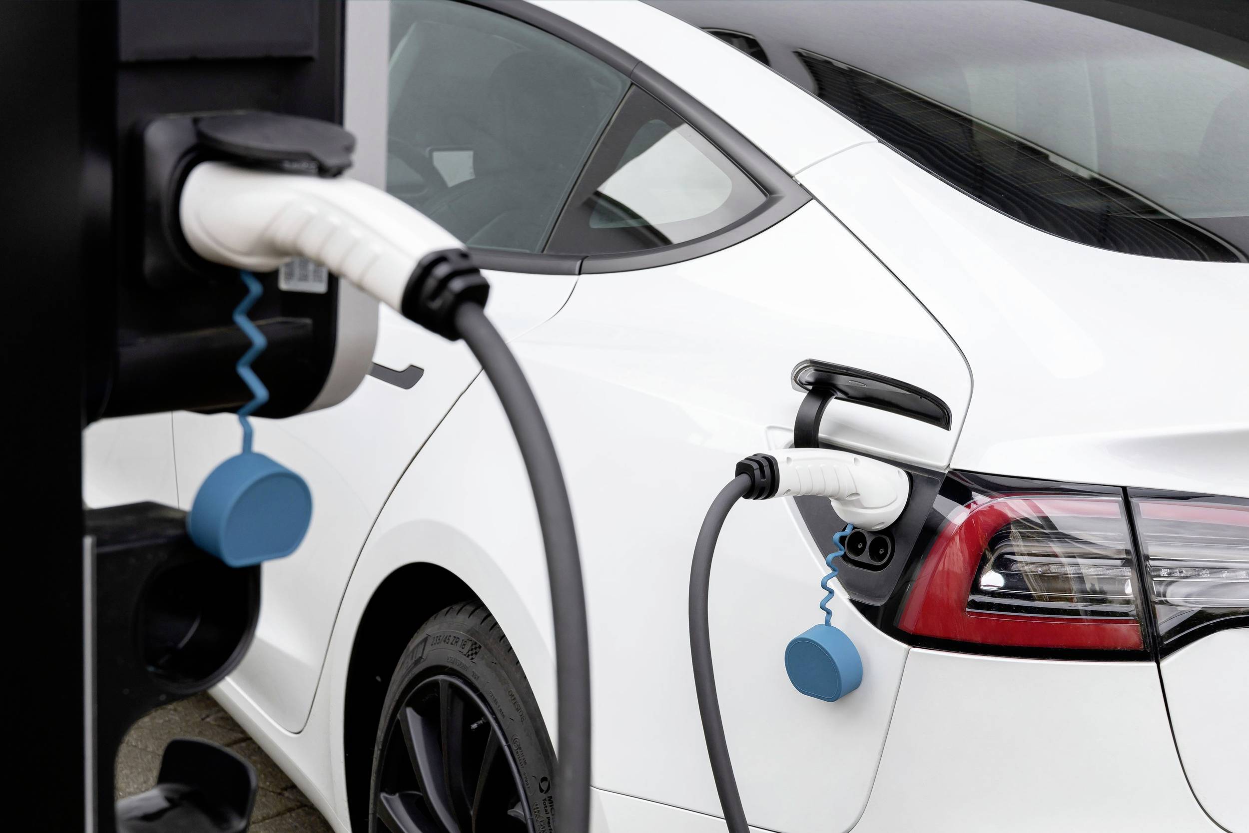 Bílý elektromobil zapojený do nabíjecí stanice. Nabíjecí kabely jsou připojeny na zadní straně vozidla.