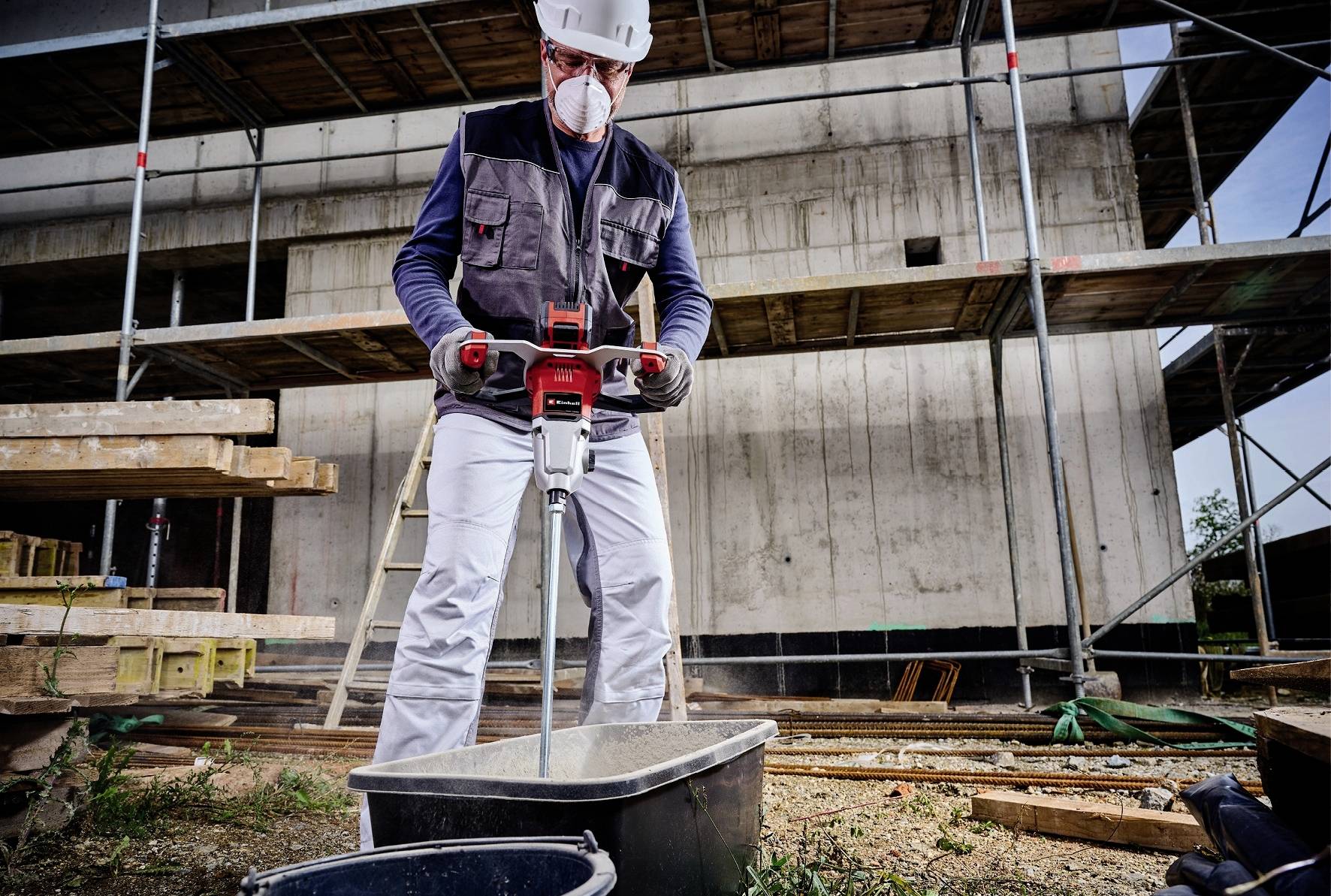 Dělník v ochranném oděvu míchá beton pomocí elektrického míchadla na staveništi před rozestavěnou budovou.