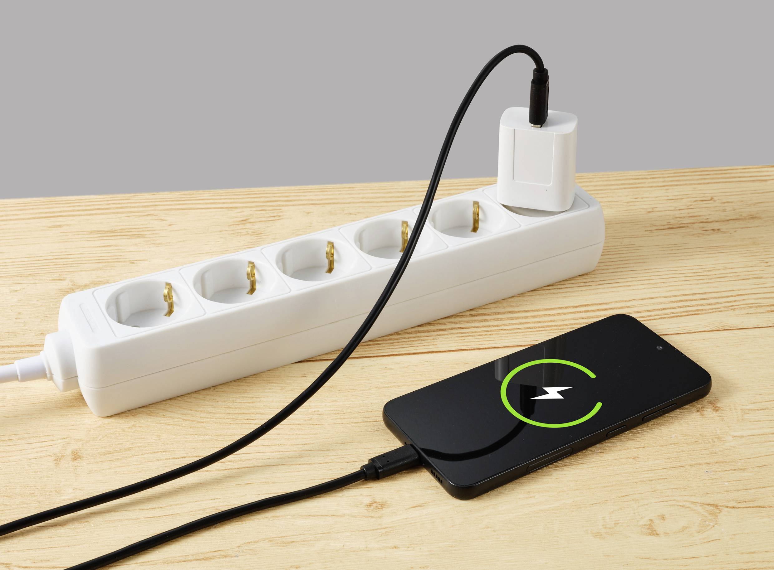 Mobilní telefon je nabíjen pomocí nabíjecího kabelu a adaptéru zapojeného do elektrické zásuvky na dřevěném stole.