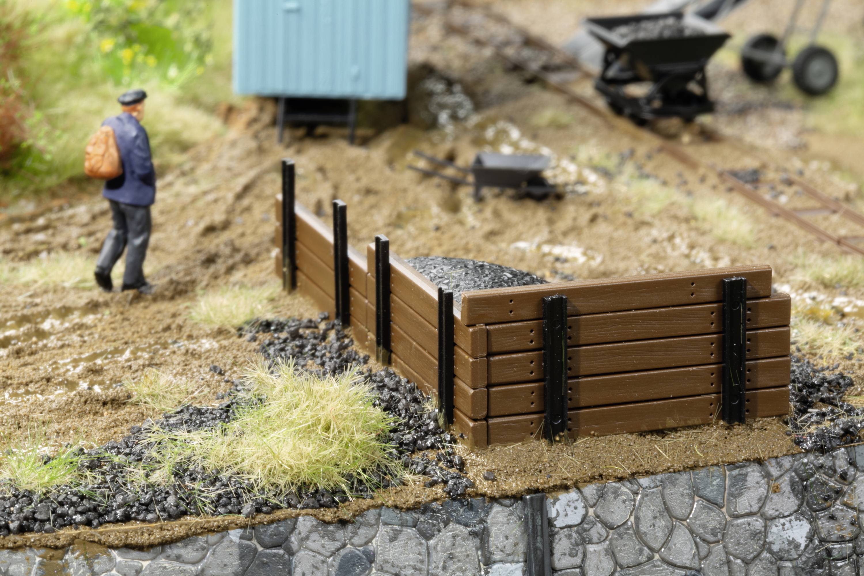Miniaturní model zobrazuje železničního dělníka s oranžovým batohem, dřevěnou nakládací rampou a kolečka na kolejích.