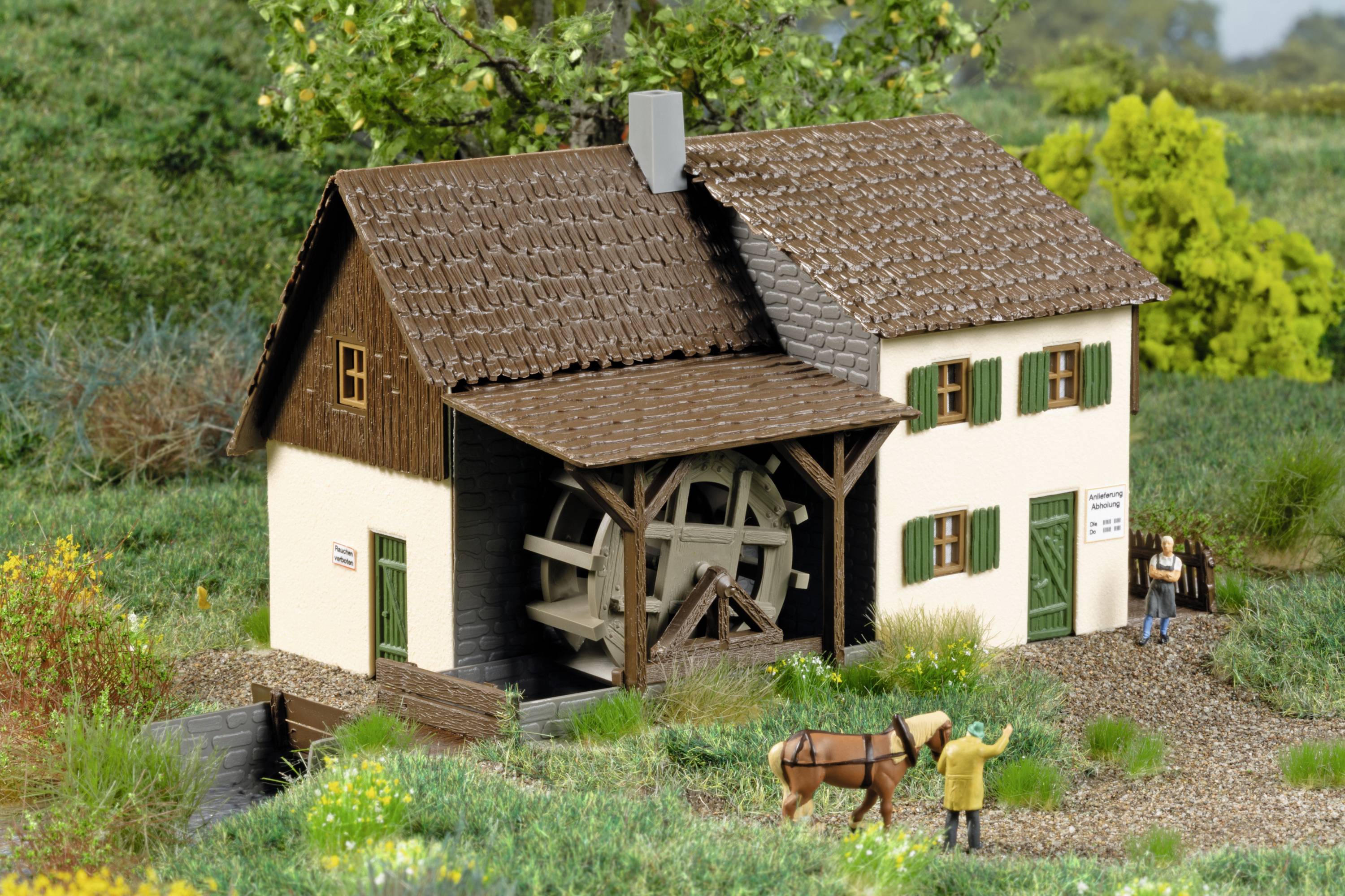 Miniaturní model vodního mlýna s otáčejícím se mlýnským kolem, obklopený zelnou krajinou. Před mlýnem stojí postava s koněm.