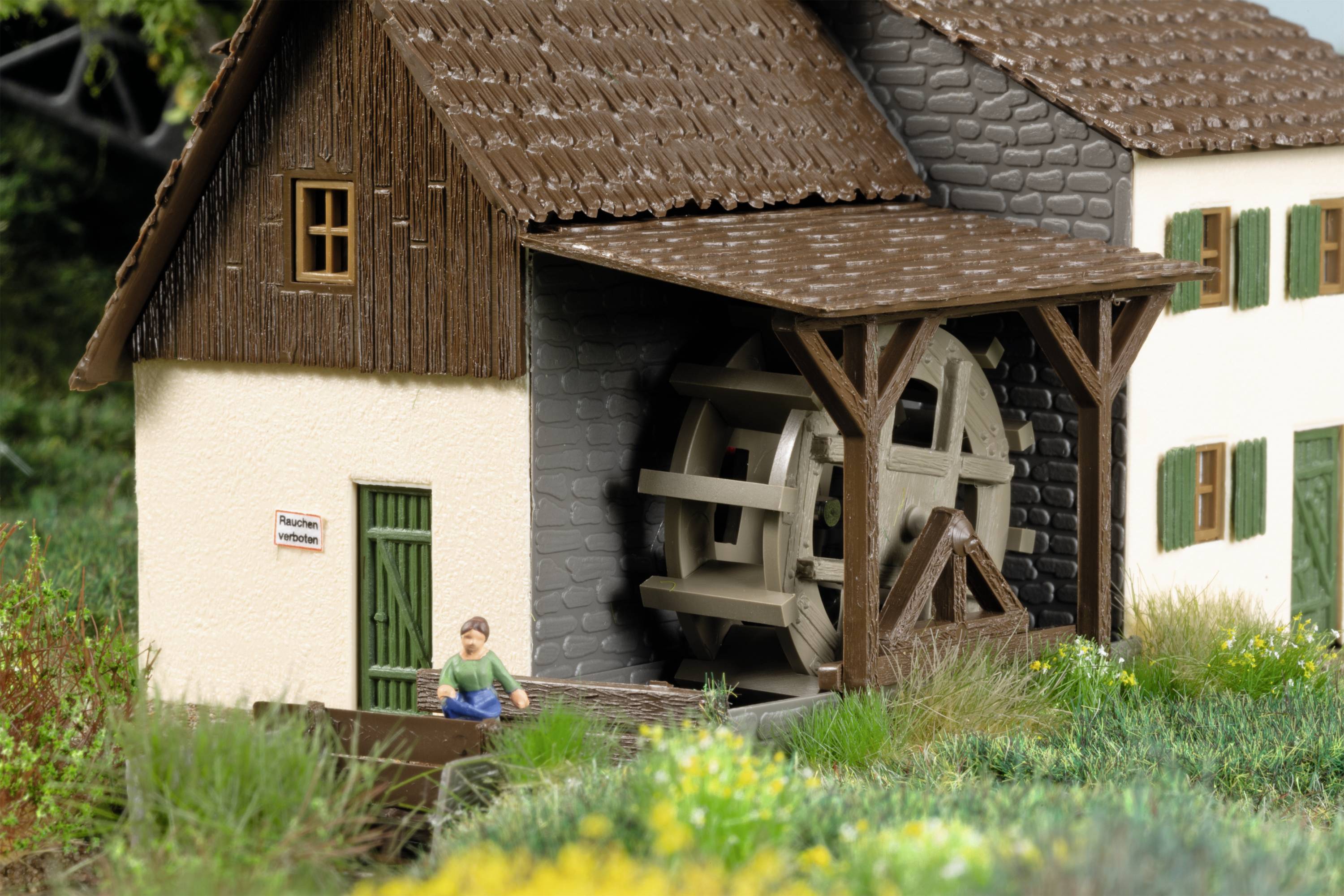 Model domu s vodním kolem. Před dřevěnou budovou sedí miniaturní figurka. Okolí porostlé trávou a květinami.