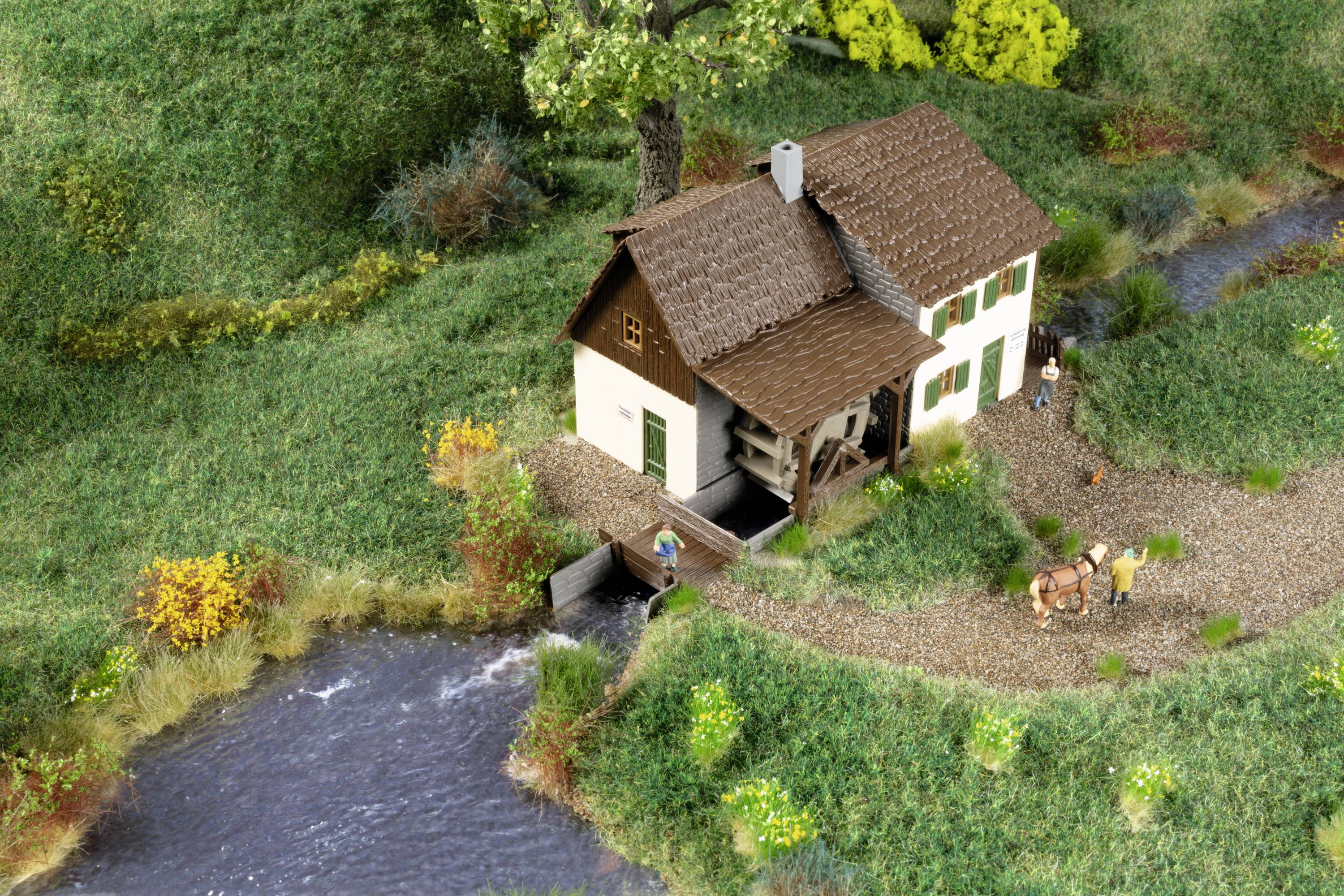 Miniaturní model venkovského domu s postavami, koněm a potokem v zelené krajině.
