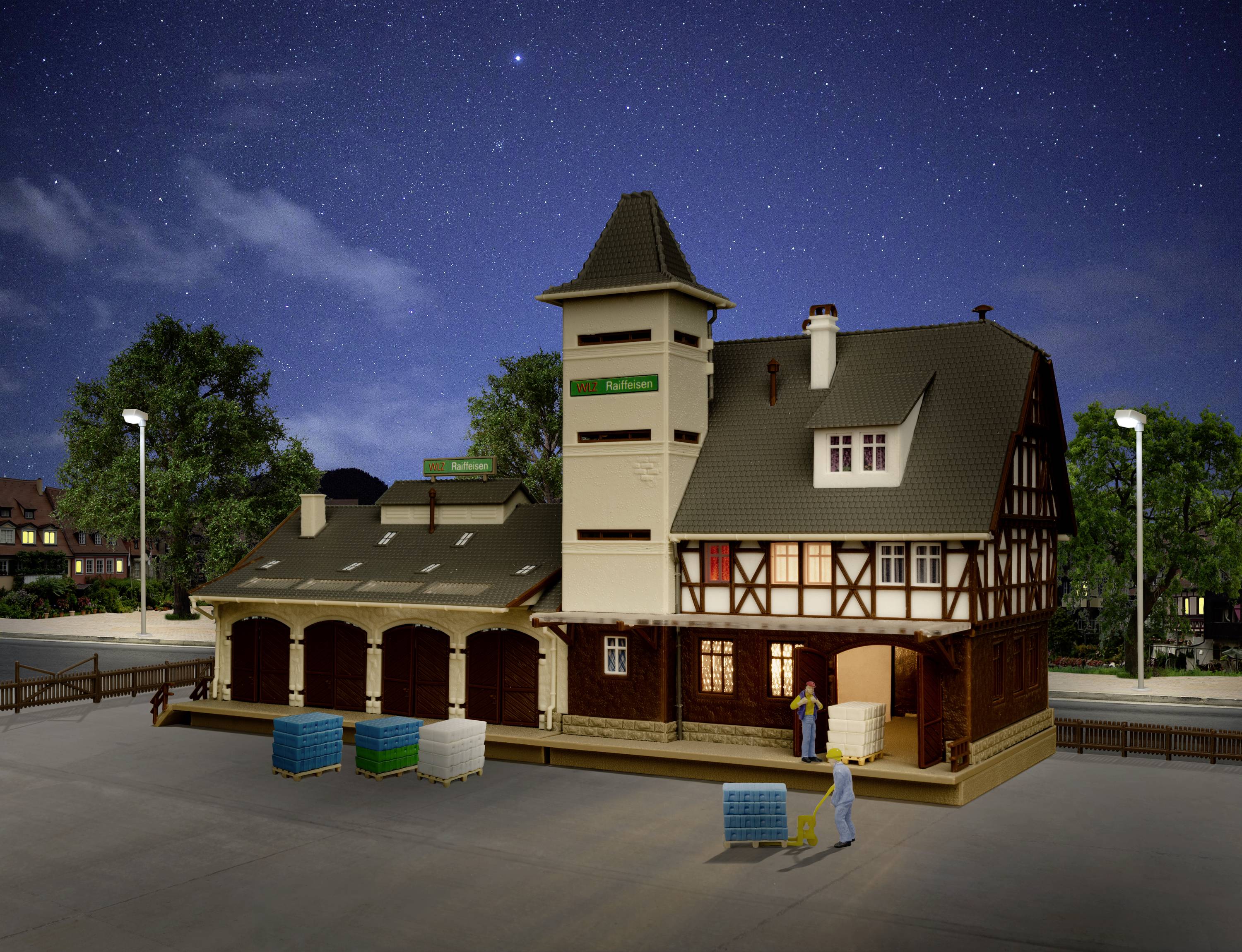 Model budovy železniční stanice v nočním osvětlení pouličních lamp, věrná historická rekonstrukce. Dvě postavy nakládají nákladovou plošinu zbožím.