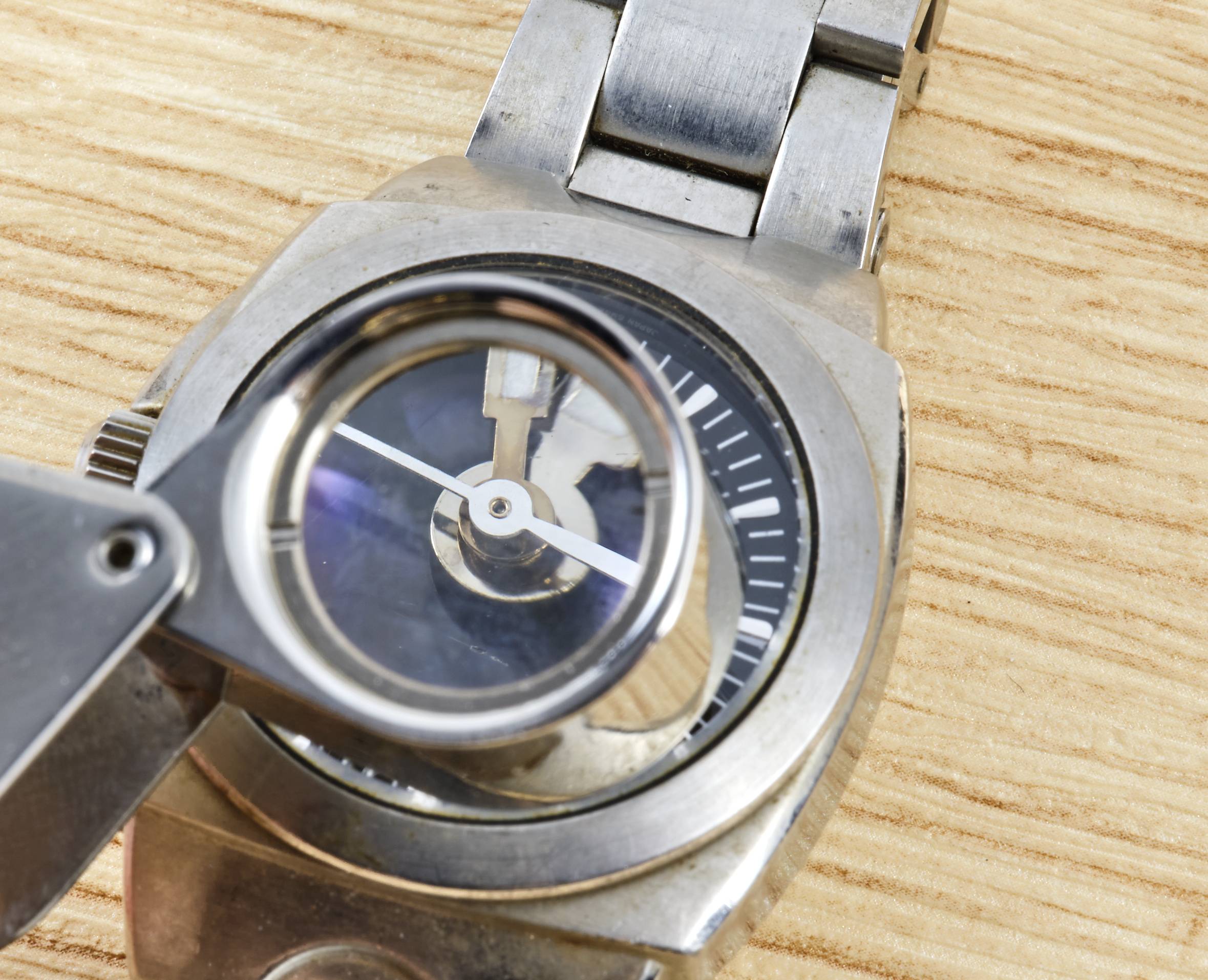 Hodinky na dřevěném povrchu jsou zkoumány lupou. Ručičky a ciferník jsou viditelné ve zvětšení.