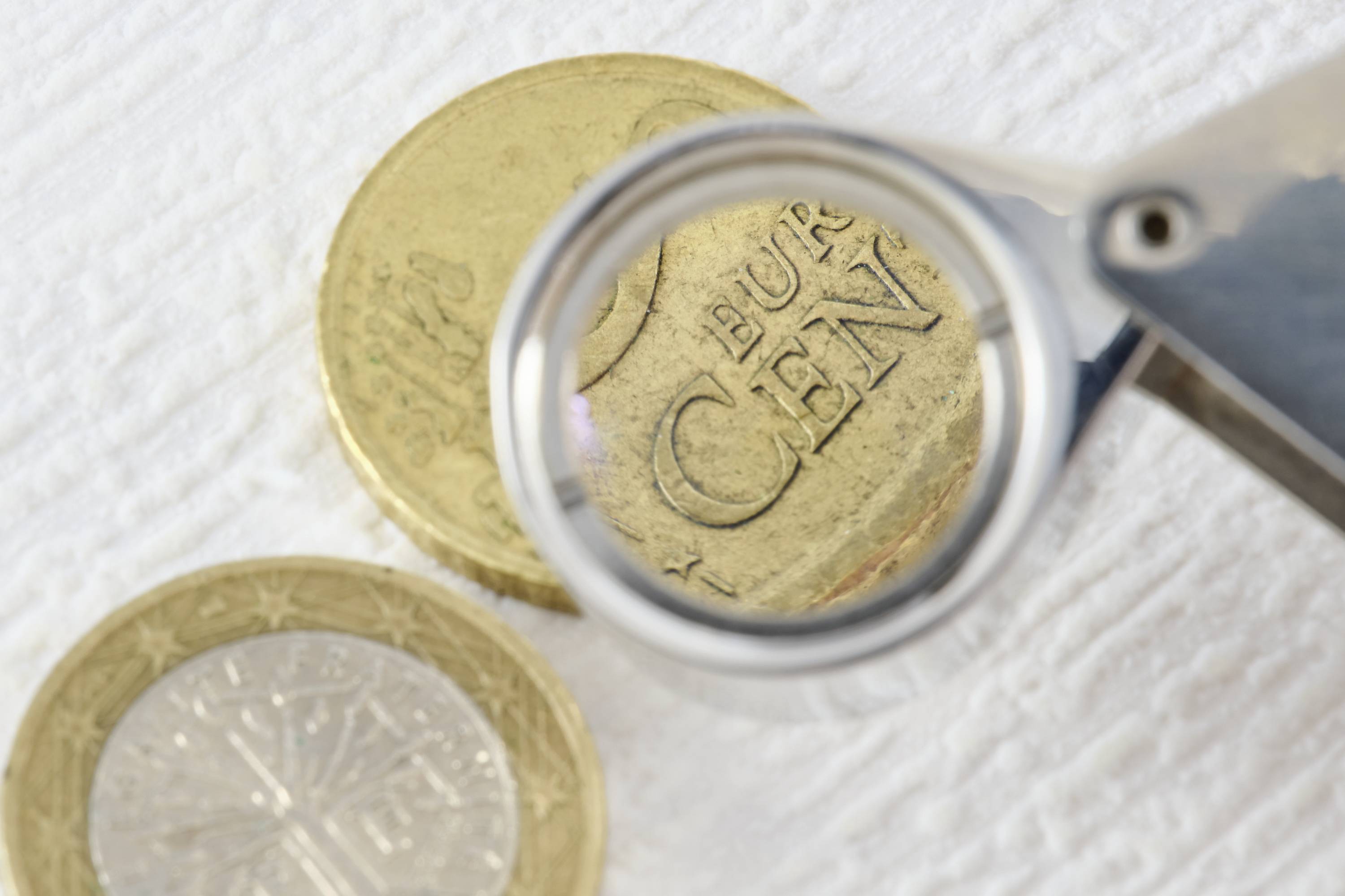 Lupa zvětšuje nápis 'EURO CENT' na eurominci. Dvě mince leží na bílém povrchu.