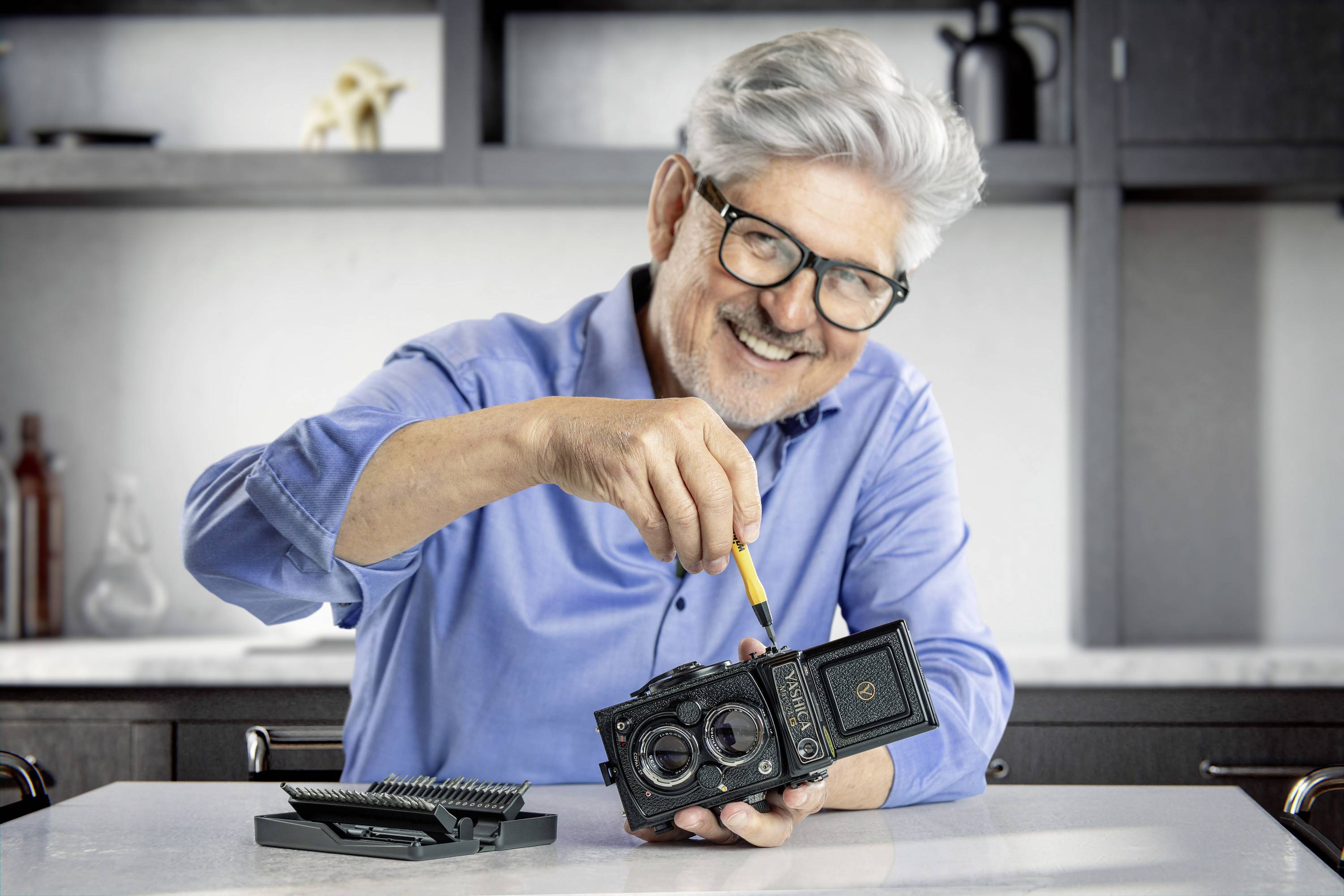Starší muž sedí u stolu a s úsměvem opravuje starý fotoaparát pomocí šroubováku. V pozadí jsou police.