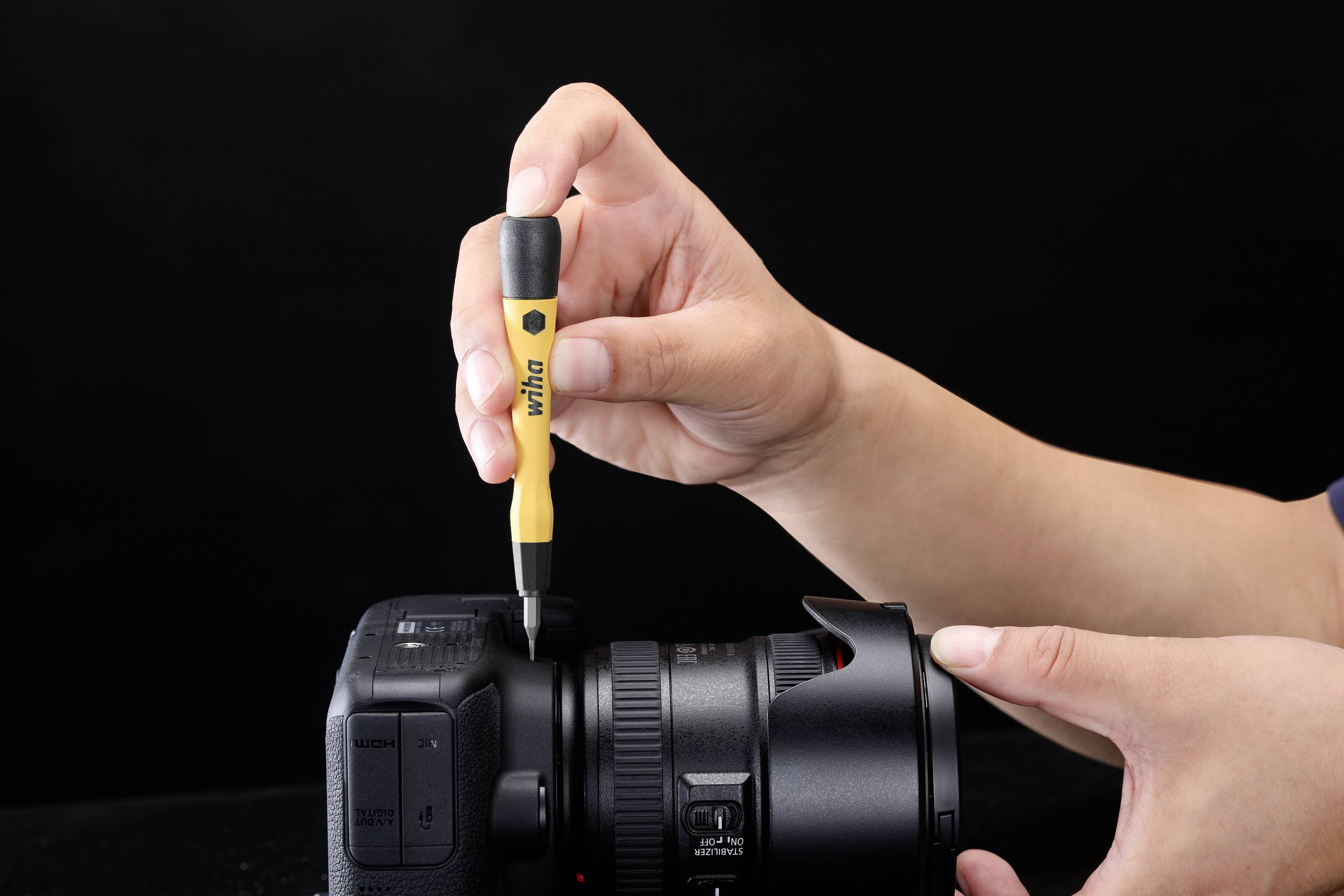 Osoba opravuje fotoaparát žlutým šroubovákem. Objektiv je upevněn a ruka drží šroubovák nad šroubem.